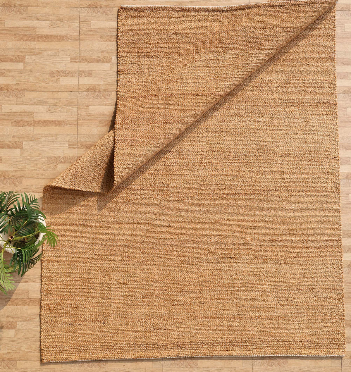 The Handmade Natural Color Hand Woven Area Rug SR-064 by RAMSHA HOME is displayed on a light wood floor, with one corner artfully folded over. A green potted plant peeks into the lower left corner, enhancing the artisanal home décor ambiance.