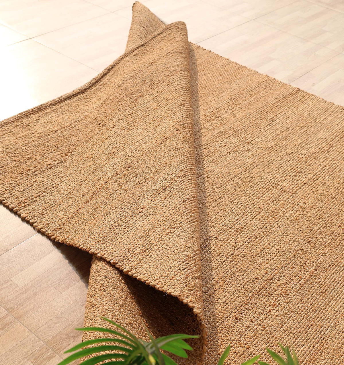 The Handmade Natural Color Hand Woven Area Rug SR-064 by RAMSHA HOME, crafted from natural fibers, is partially folded on a light wooden floor, introducing an artisanal touch to the décor. A small green plant appears in the lower left corner of the image.
