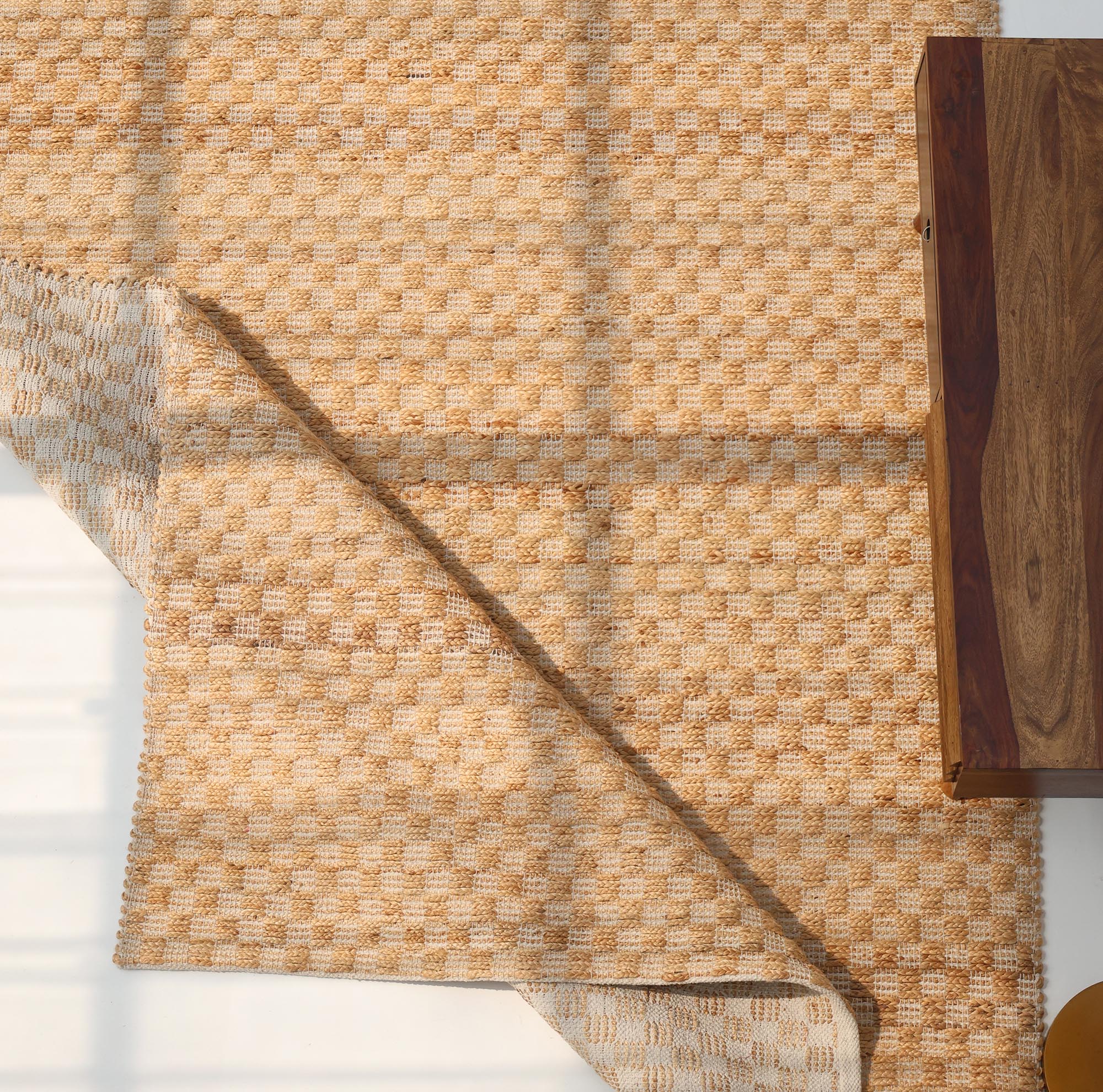 The Handmade Natural Color Hand Woven Area Rug SR-065 by RAMSHA HOME is partly folded at one corner on a light floor beside a wooden table, with soft sunlight casting shadows across this cozy, artisanal home décor scene.