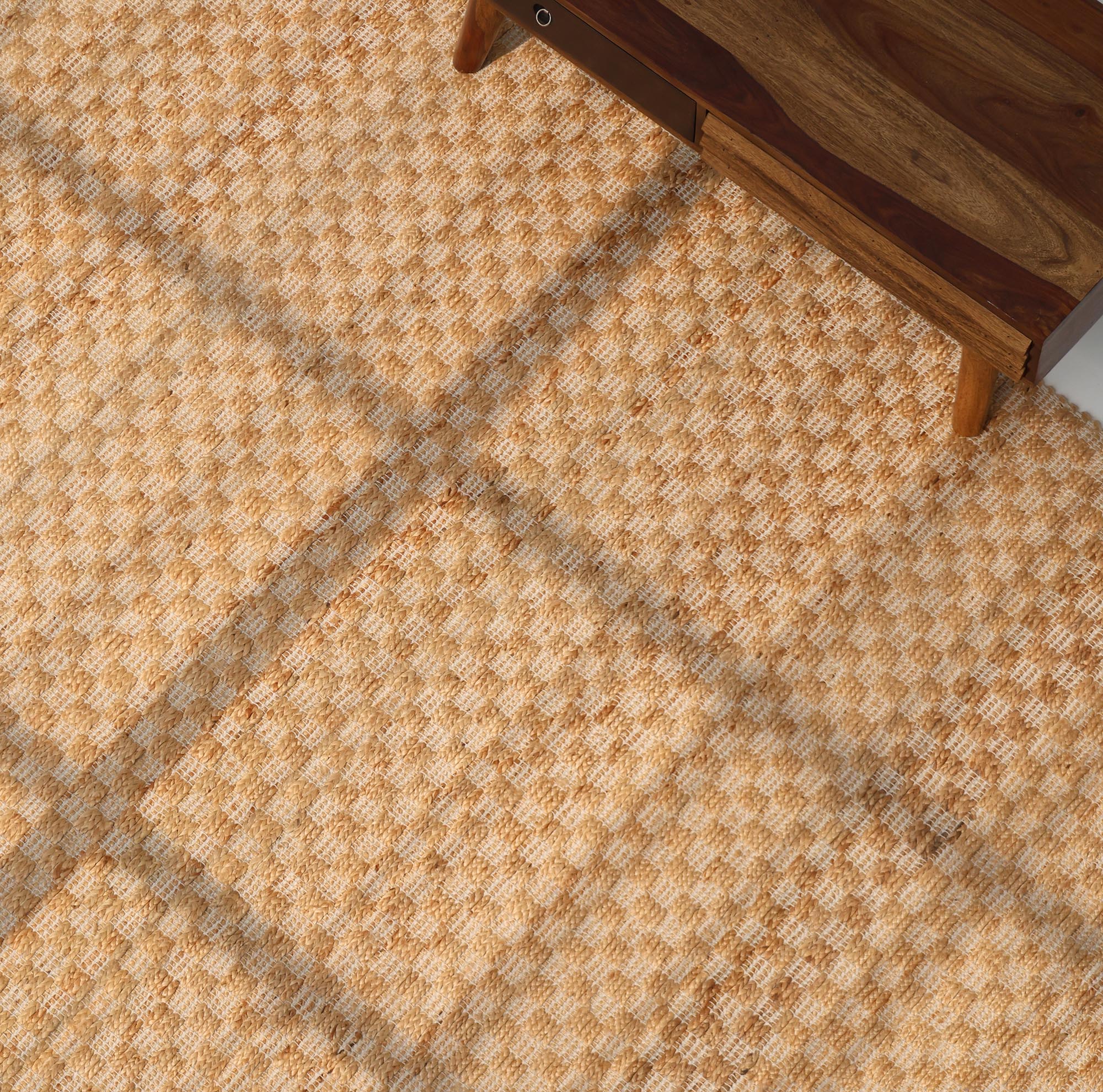 A close-up of the RAMSHA HOME Handmade Natural Color Hand Woven Area Rug SR-065, featuring a textured beige and light brown checkered pattern, partially placed beneath wooden furniture with angled legs.