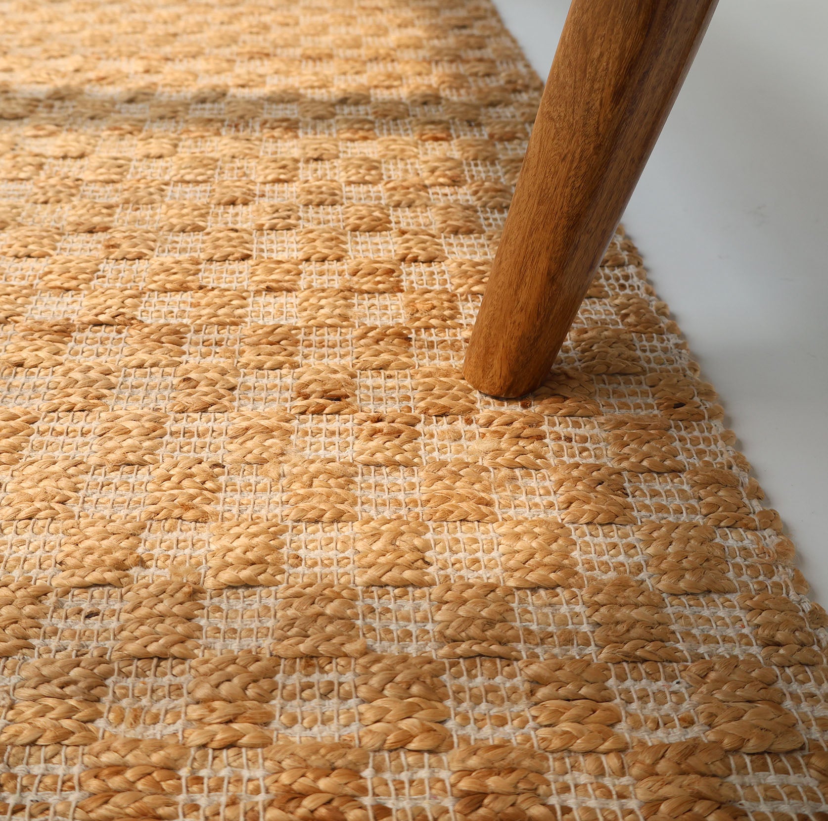 Close-up of the RAMSHA HOME Handmade Natural Color Hand Woven Area Rug SR-065 in beige and tan checks, with a wooden furniture leg on it, showcasing its textured pattern and artisanal style.