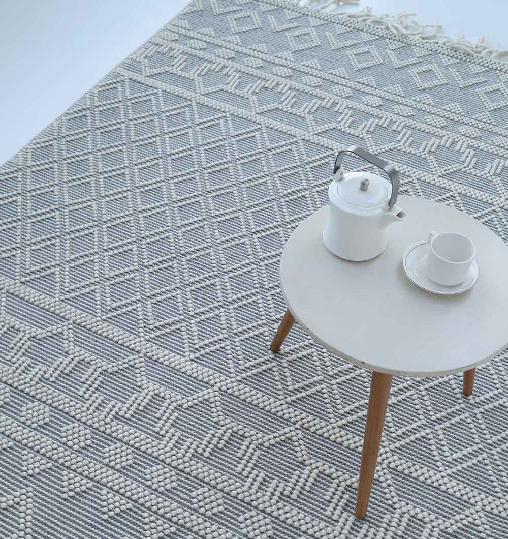 A round white table with a teapot and cup sits on the RAMSHA HOME Wool & Cotton Woven Rug Area Rug SRN-004, which features textured gray and white geometric patterns and fringe along one edge.