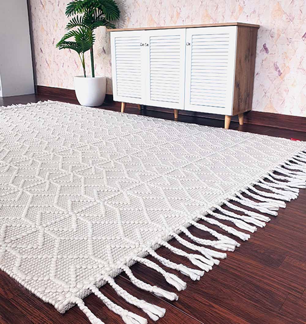 The RAMSHA HOME Handmade Woven Rug in natural white (SRN-007) with fringed edges is laid on a dark wooden floor before a white cabinet, while a potted plant sits beside the cabinet against a patterned wall.