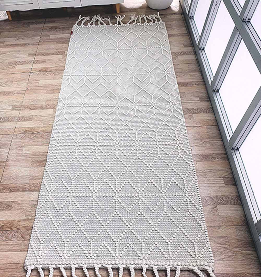 The RAMSHA HOME Handmade Woven Rug Natural White Color Fringes Decor Rug SRN-007, featuring a diamond pattern and fringed ends, adds texture and brightness to the wooden floor near the windowed wall.