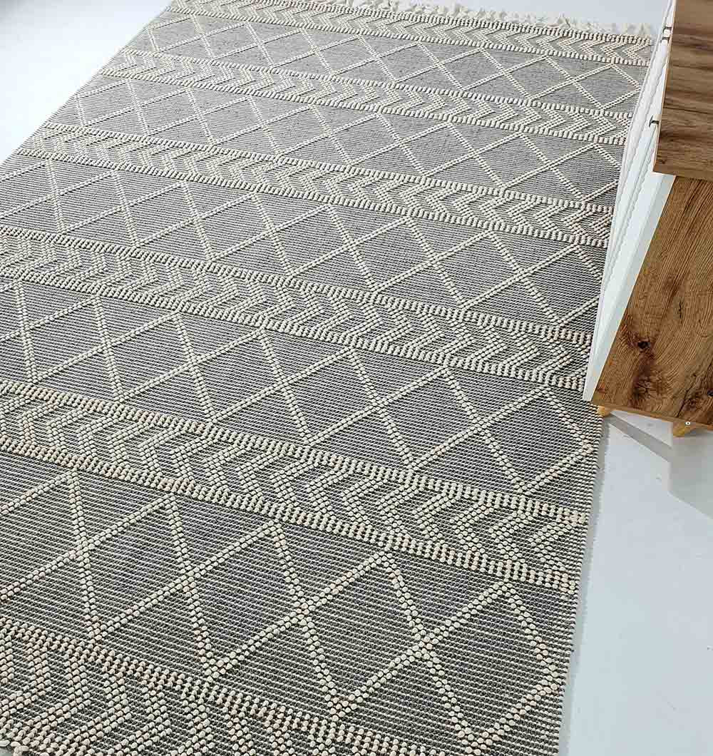 The RAMSHA HOME Ramadan Prayer Rugs Natural White Floor Woven Rug with Fringes SRN 020, featuring gray and white geometric patterns, is displayed on a light floor beside a wooden surface.