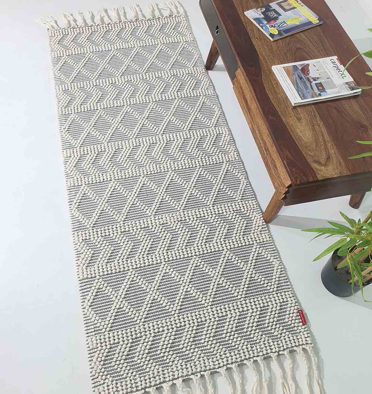 The RAMSHA HOME Ramadan Prayer Rugs Natural White Floor Woven Rug with Fringes SRN 020, featuring a geometric pattern, is placed beside a wooden coffee table with magazines. Green plant leaves are seen in the corner.
