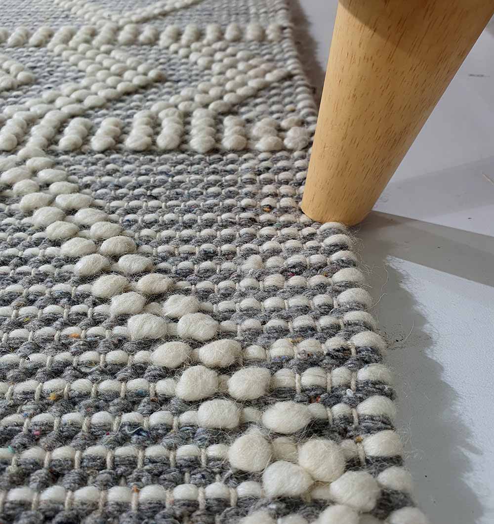 Close-up of the RAMSHA HOME Ramadan Prayer Rugs Natural White Floor Woven Rug with Fringes SRN 020, featuring raised white patterns on a gray base beside wooden furniture on a light floor.