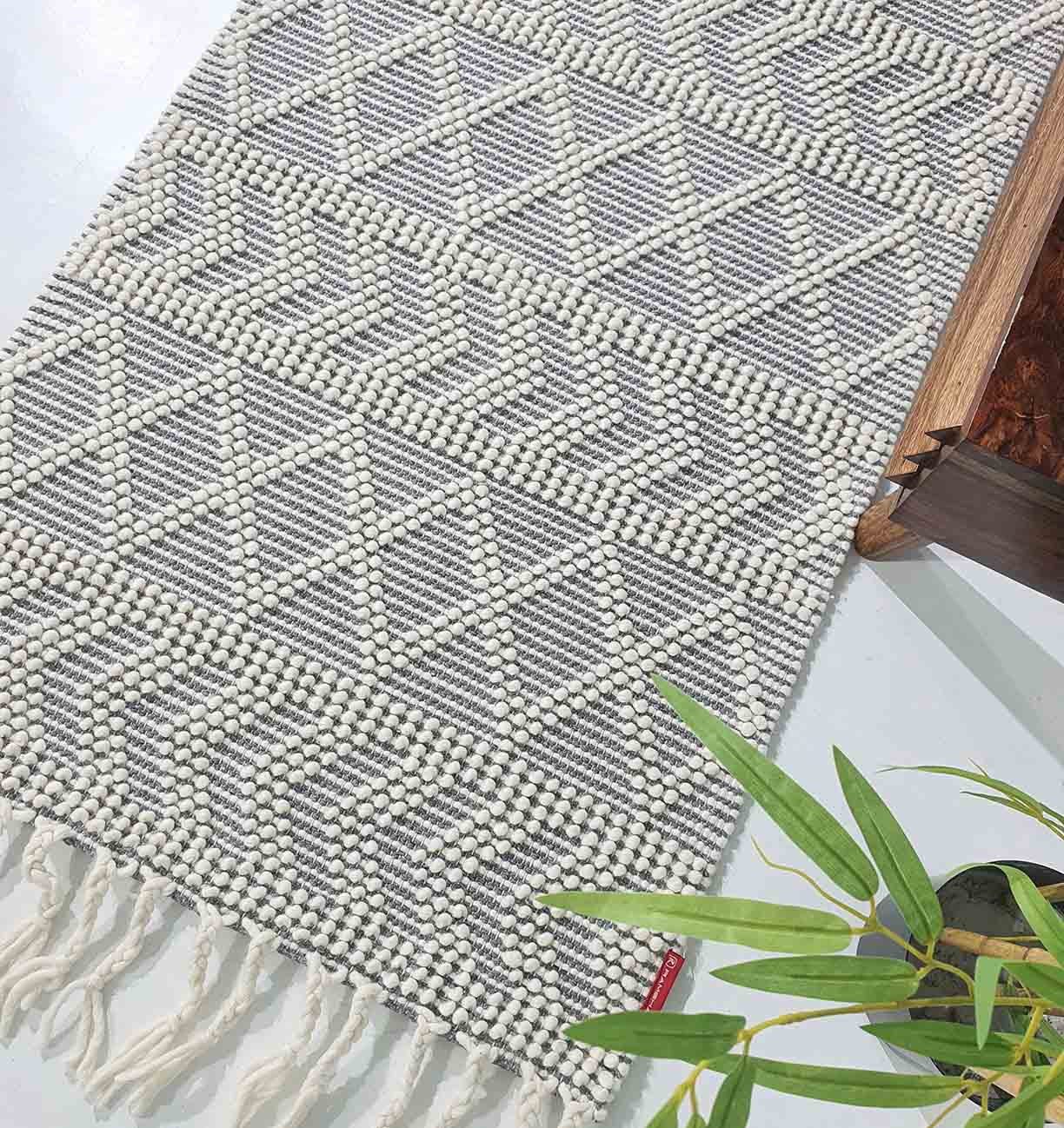 The RAMSHA HOME Ramadan Prayer Rugs Natural White Floor Woven Rug with Fringes SRN 020, featuring a geometric diamond pattern in white and gray, is displayed next to a wooden surface and a green plant.