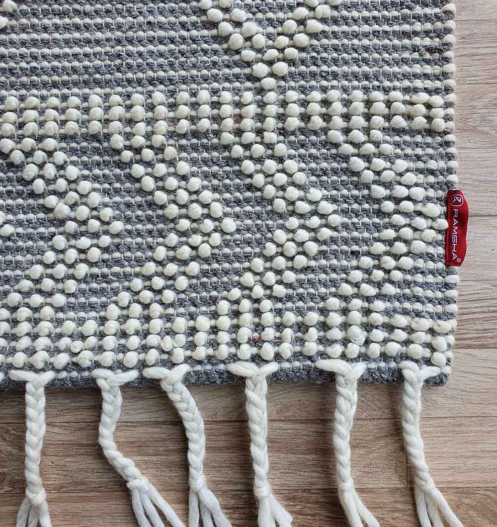 A close-up of the RAMSHA HOME Ramadan Prayer Rugs Natural White Floor Woven Rug with gray and white geometric patterns, textured dots, fringes on the edge, and a red fabric tag, displayed on a wooden floor.