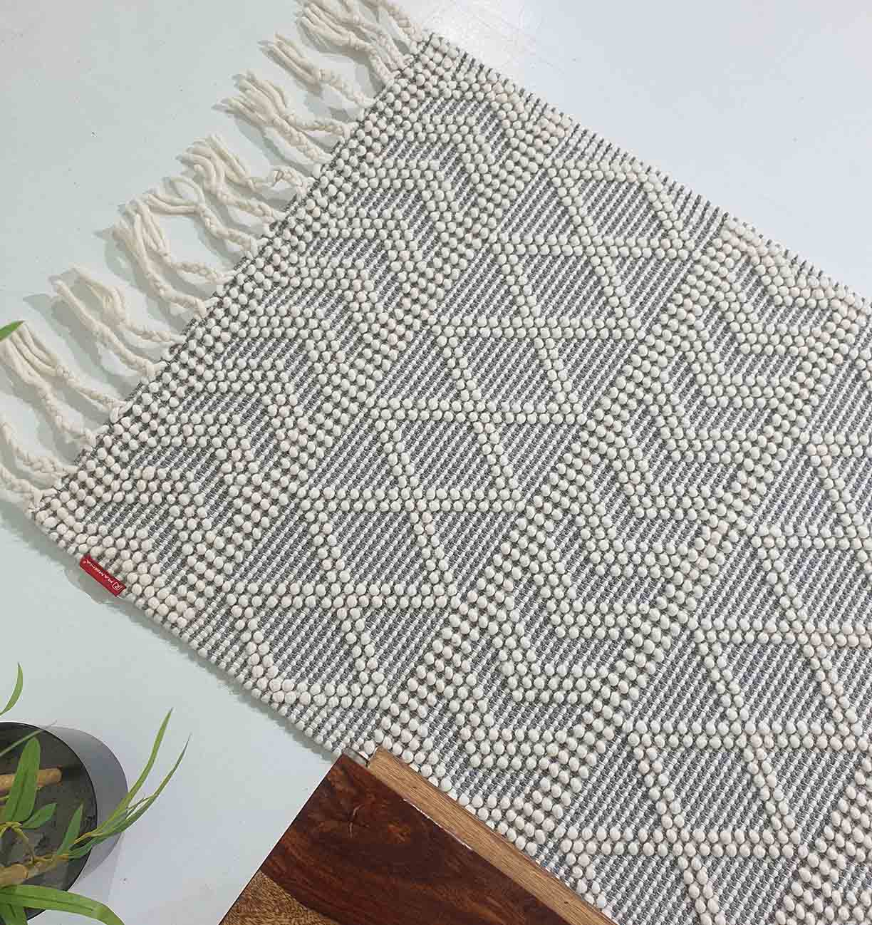 The RAMSHA HOME Ramadan Prayer Rugs Natural White Floor Woven Rug with Fringes SRN 020 features a geometric diamond and chevron pattern in white and gray, finished with white fringe on one end. A plant and wooden table are seen nearby.