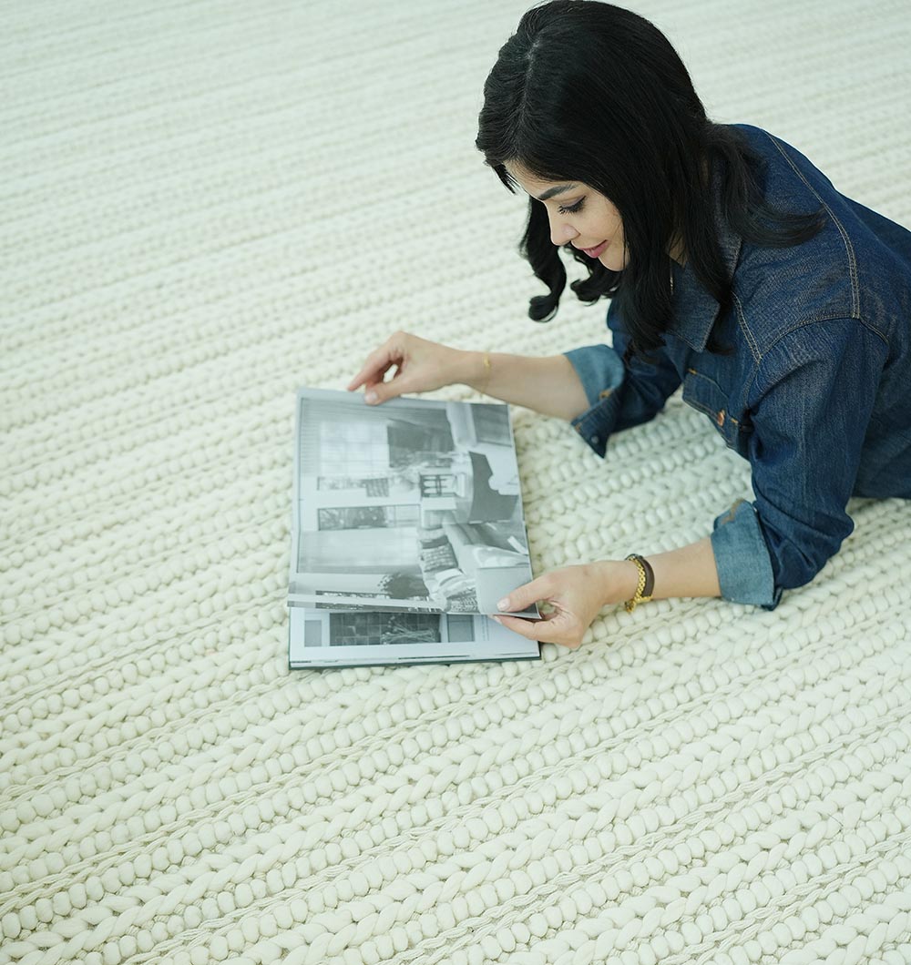 A woman with dark hair, in a blue shirt, relaxes on the RAMSHA HOME Natural White Woven Rug For Home Decore SRN-028 while viewing a black-and-white photo book.