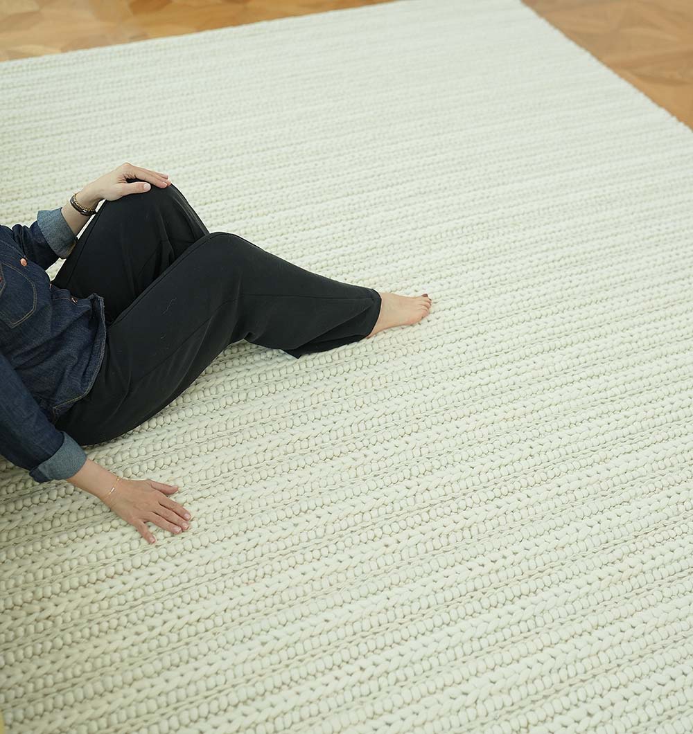 A person sits barefoot on the RAMSHA HOME Natural White Woven Rug For Home Decore SRN-028, with just their lower body and hand visible, wearing dark pants and a denim shirt on a wooden floor.