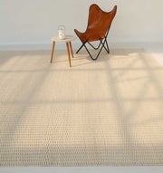A minimalist room features the RAMSHA HOME Handmade Natural White Woven Rug SRN-030, a brown leather butterfly chair, and a small round wooden side table with a glass teapot and cup. Soft sunlight casts gentle shadows across the floor.