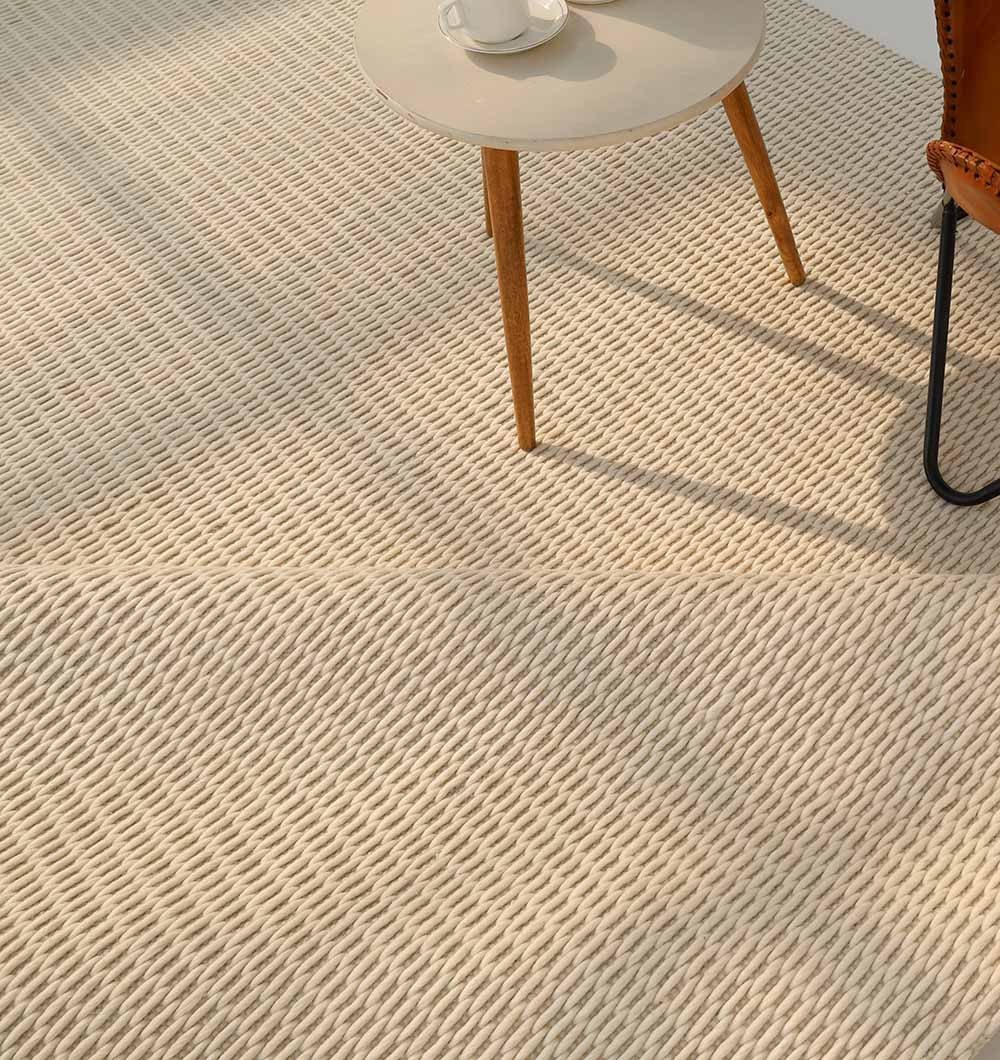 The Handmade Natural White Woven Rug For Home Decor SRN-030 by RAMSHA HOME lies beneath a round, light-hued table with wooden legs and part of a brown leather chair, as sunlight casts gentle shadows across the rug.