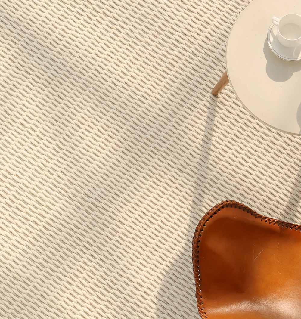 A close-up of the RAMSHA HOME Handmade Natural White Woven Rug For Home Decor SRN-030, with a round beige table holding a white cup and saucer beside a brown leather chair, as sunlight casts soft shadows across the scene.