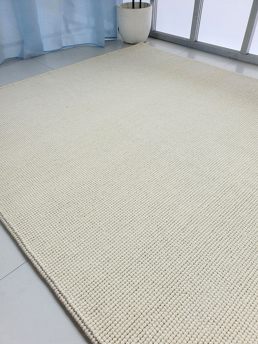 The Solid Loop Pile Woven Rug For Home Decor SRN-032, a large beige wool rug with textured design, sits on a white tile floor near a window with light blue curtains and a white potted plant in the corner.