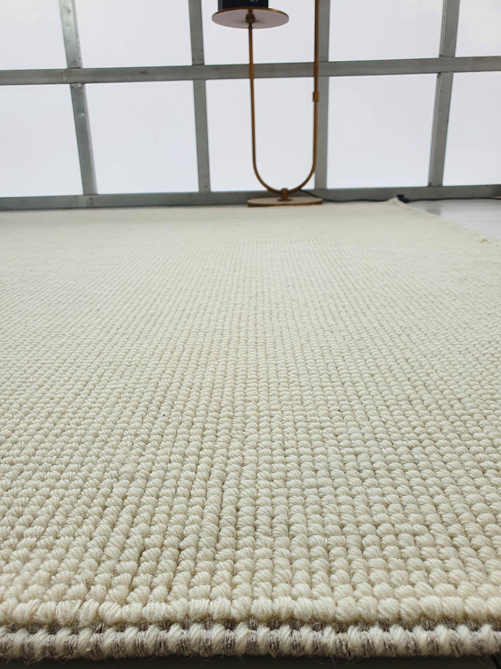 Close-up of the Solid Loop Pile Woven Rug For Home Decor SRN-032 in beige, featuring a textured surface on the floor with a modern gold lamp and large windows in the background letting in natural light.