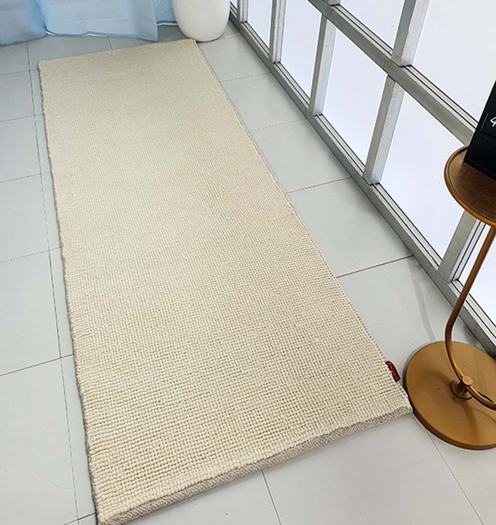 The Solid Loop Pile Woven Rug For Home Decor SRN-032, a long beige flatweave rug, is laid on white tiled flooring near frosted glass windows. A small round golden side table and a white spherical decor piece complete the setting.
