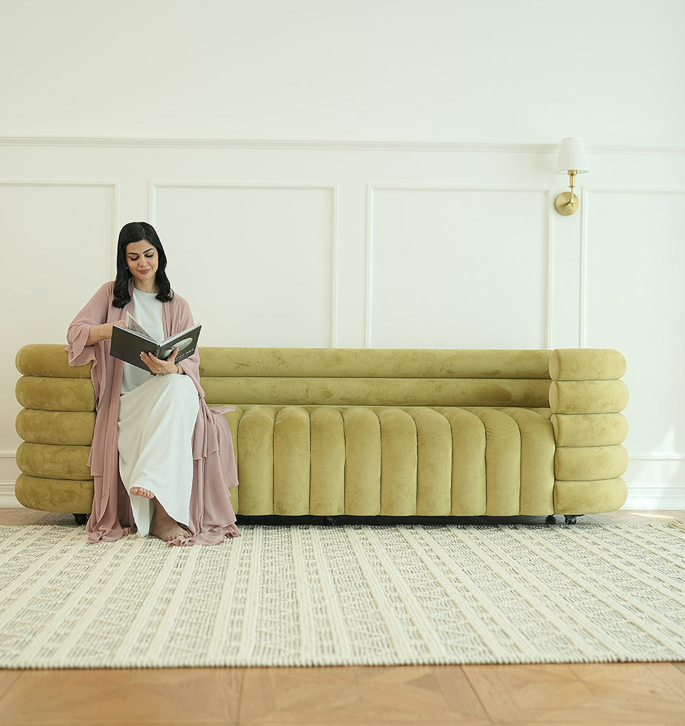 A woman in a light dress and mauve robe reads on a modern olive-green sofa. The room features white paneled walls, a wall sconce, and is cozy with the RAMSHA HOME Wool Woven Rug Area Rug with Natural Color SRN-040 on the wooden floor.