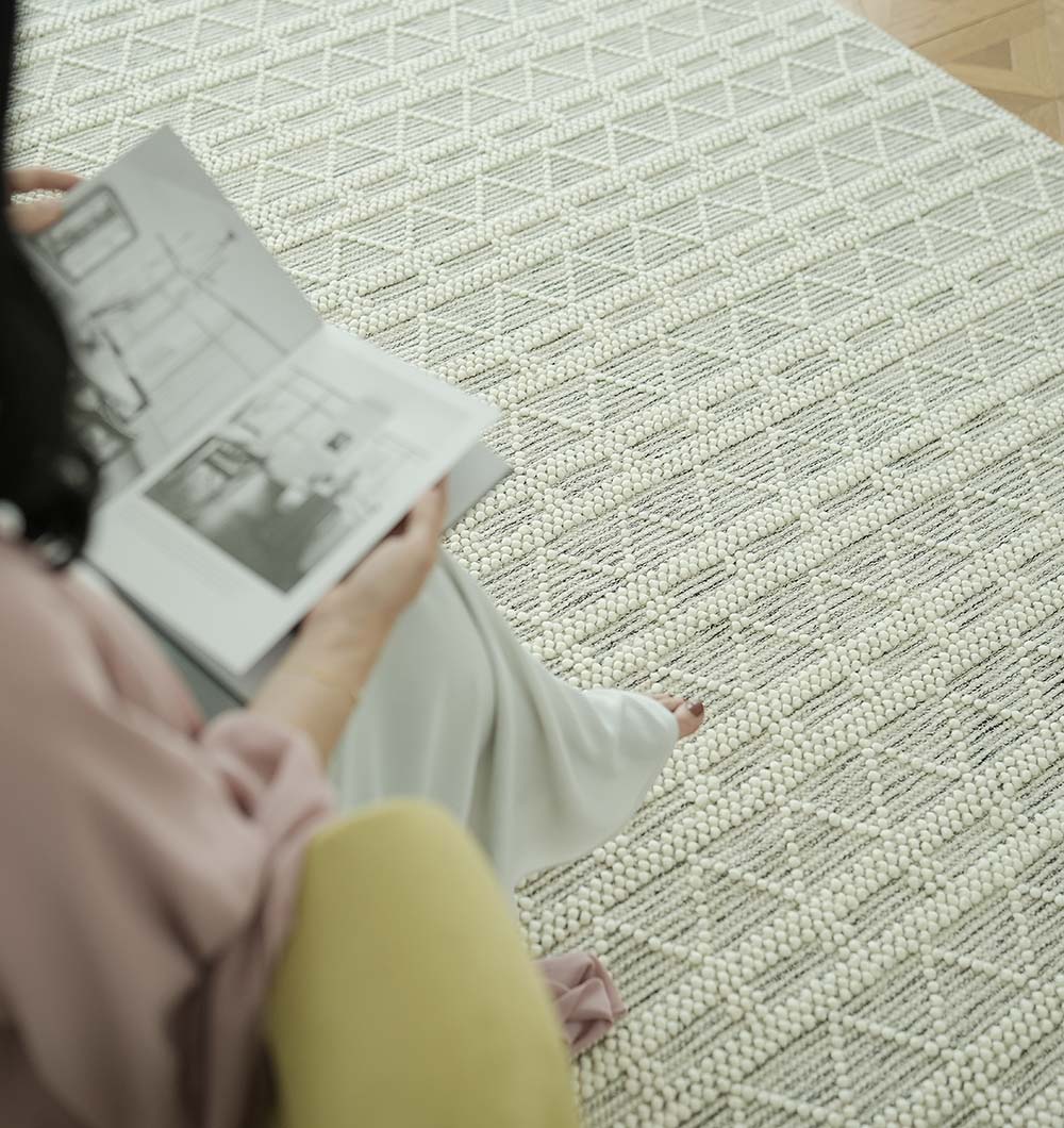 A person in light clothing reads a magazine on a chair, highlighting the RAMSHA HOME Wool Woven Rug Area Rug with Natural Color SRN-040—its textured, geometric cream and gray pattern elevates the living room’s style.