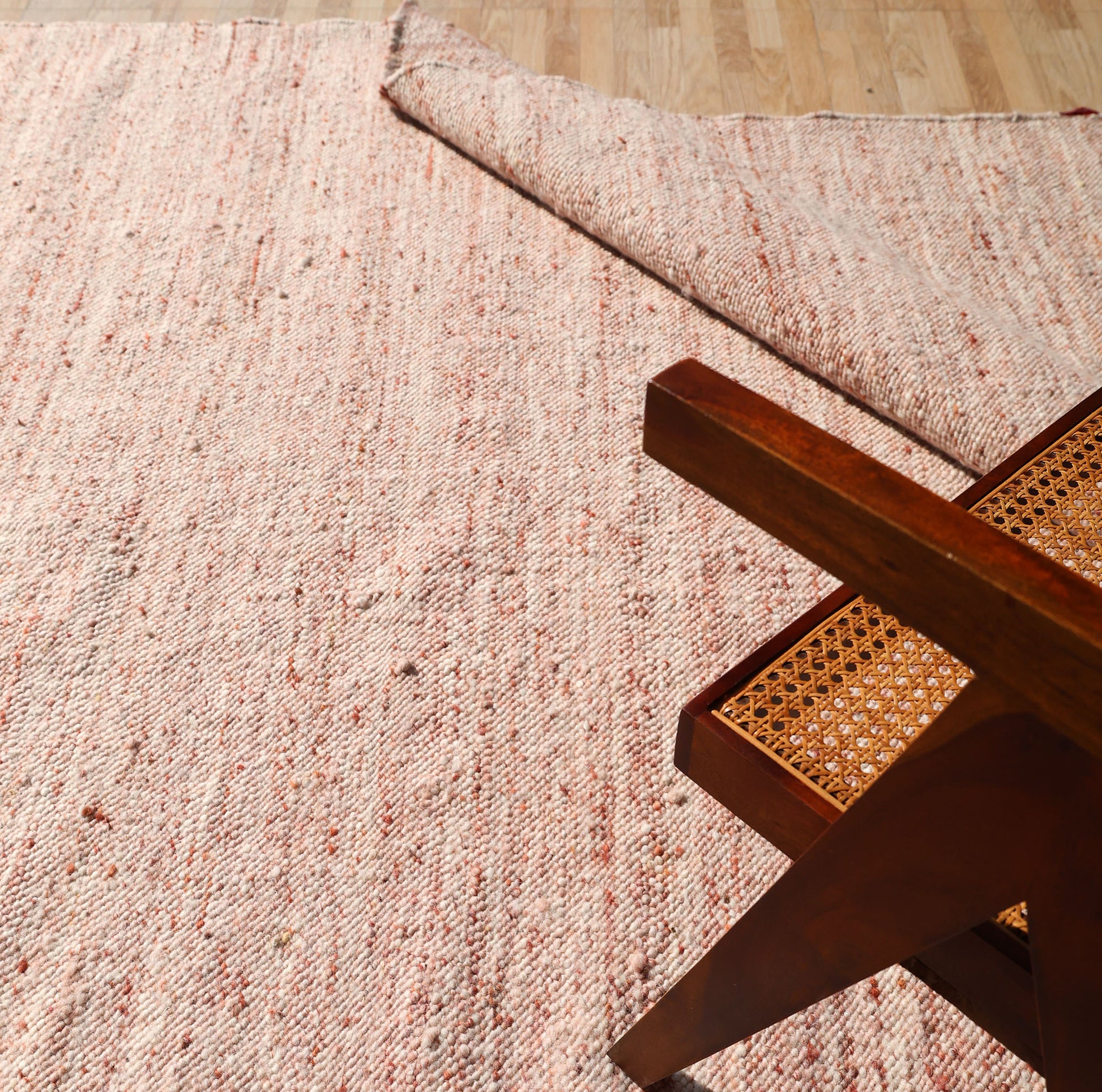 A close-up of RAMSHA HOME's Dusty Rose Artisan Flatweave Wool Rug-SRN-042 shows its light pink, textured surface with a folded corner and part of a cane-seated wooden chair on the neutral rug atop a light wood floor.