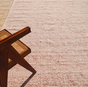 A wooden chair with a woven cane seat rests on the RAMSHA HOME Dusty Rose Artisan Flatweave Wool Rug (SRN-042), as sunlight highlights its neutral tones and textured pink surface, showcasing traditional craftsmanship.