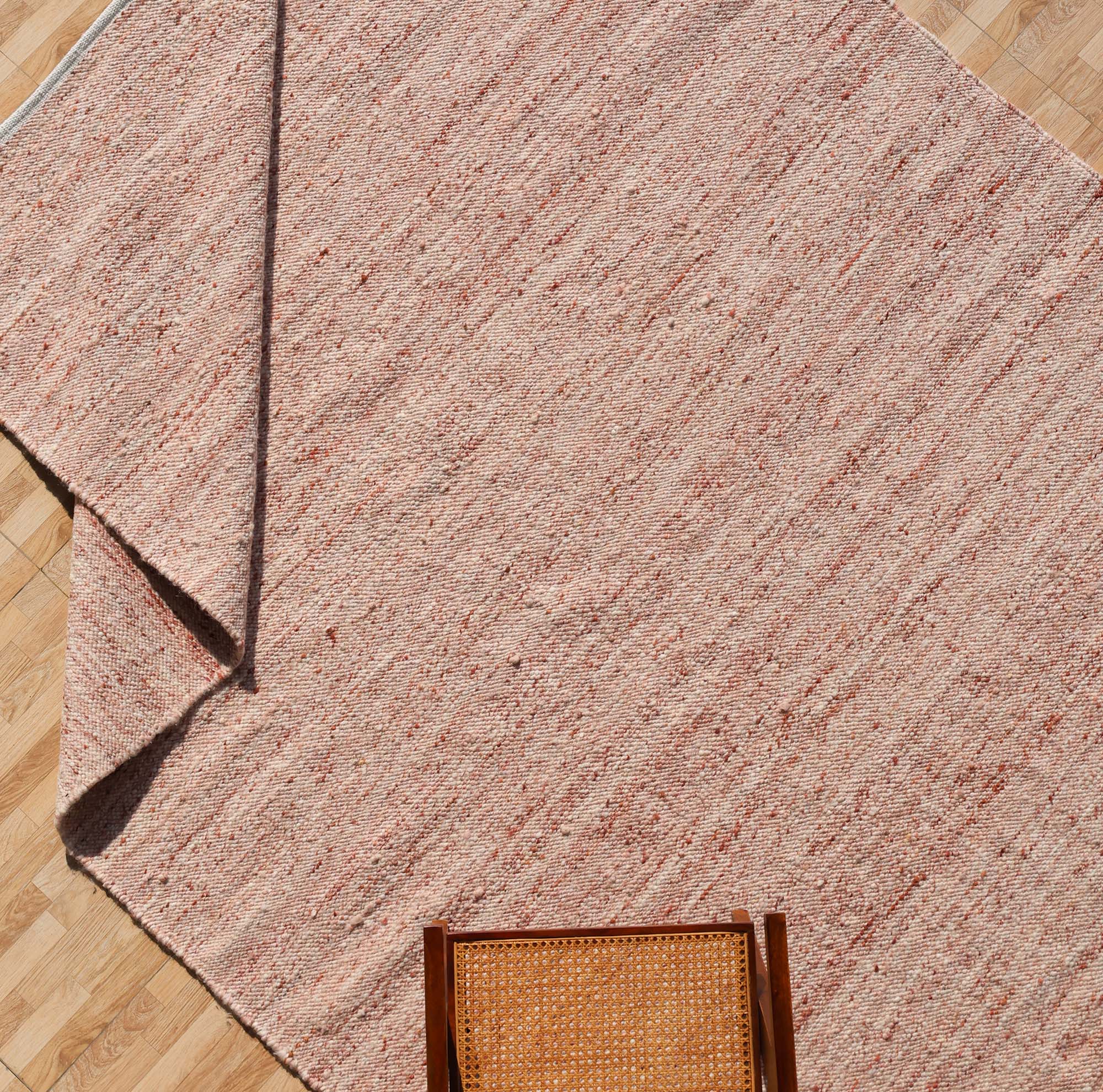 The RAMSHA HOME Dusty Rose Artisan Flatweave Wool Rug (SRN-042) is displayed on a light wood floor with a folded corner. A wooden chair with a cane seat appears in the lower right, emphasizing the rug’s neutral tones and handcrafted design.