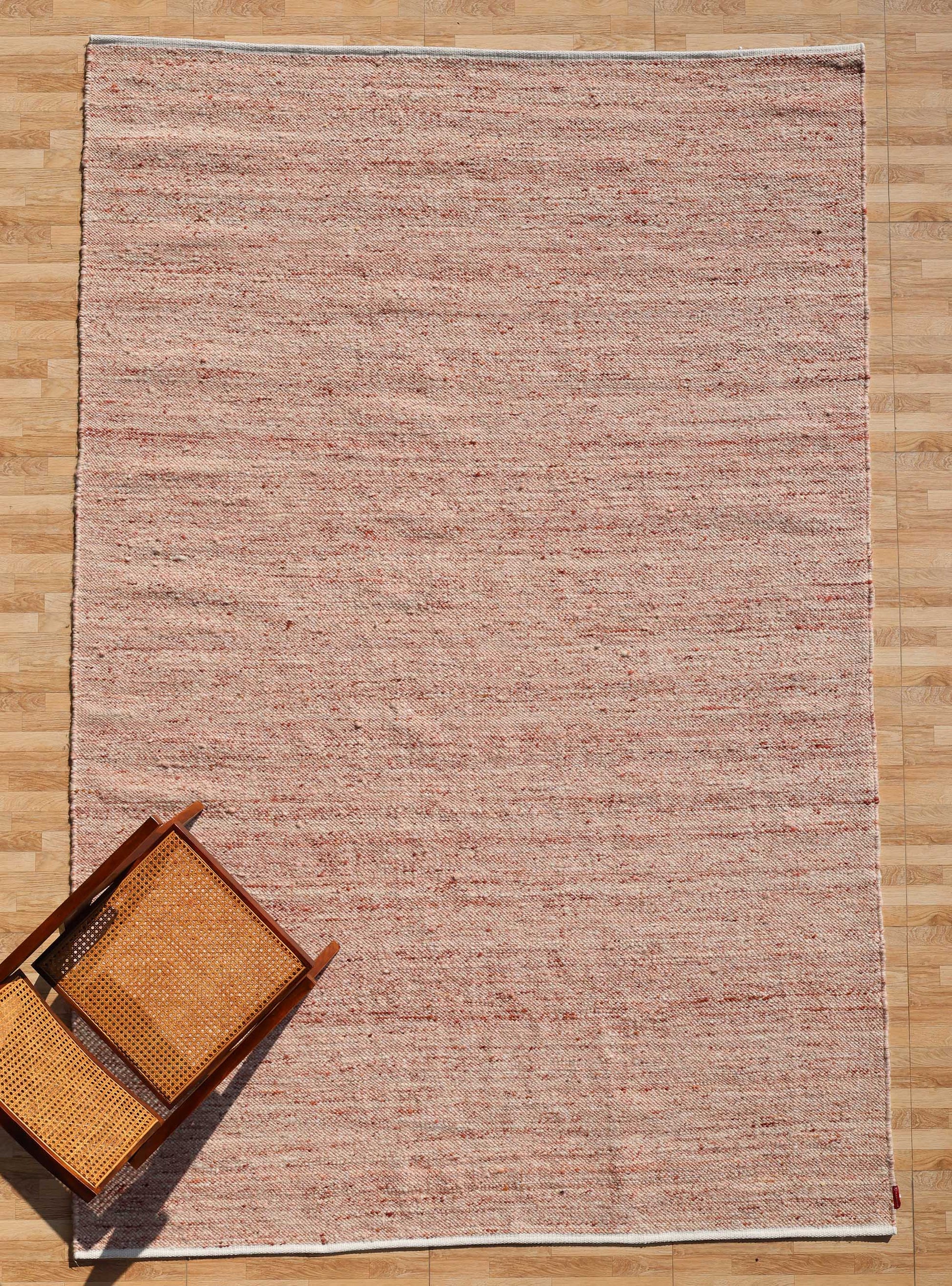 The RAMSHA HOME Dusty Rose Artisan Flatweave Wool Rug (SRN-042), featuring subtle red and beige patterns, is displayed on a wooden floor beside an overturned wooden chair with a woven seat and backrest.