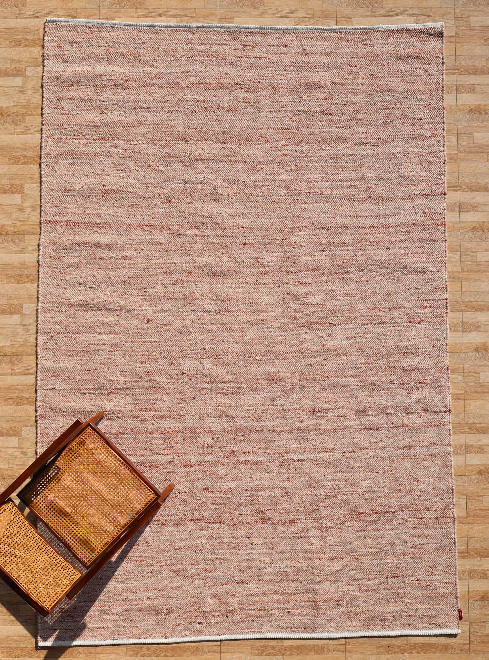 The RAMSHA HOME Dusty Rose Artisan Flatweave Wool Rug (SRN-042), featuring subtle red and beige patterns, is displayed on a wooden floor beside an overturned wooden chair with a woven seat and backrest.