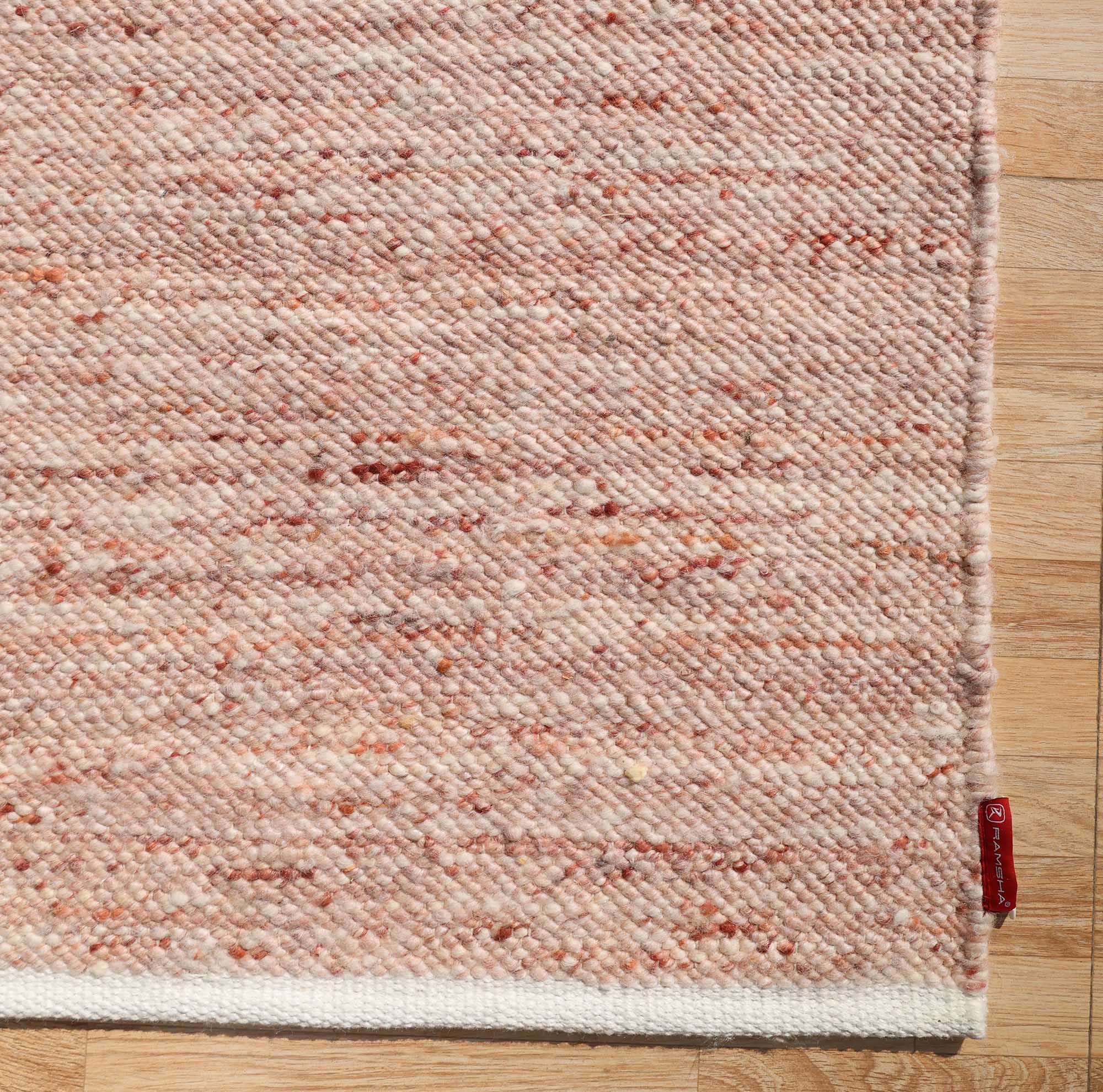 The RAMSHA HOME Dusty Rose Artisan Flatweave Wool Rug (SRN-042) features a textured light pink hue with a white border, neutral colorway, and traditional craftsmanship. A small red fabric tag adorns the corner of this handmade rug.