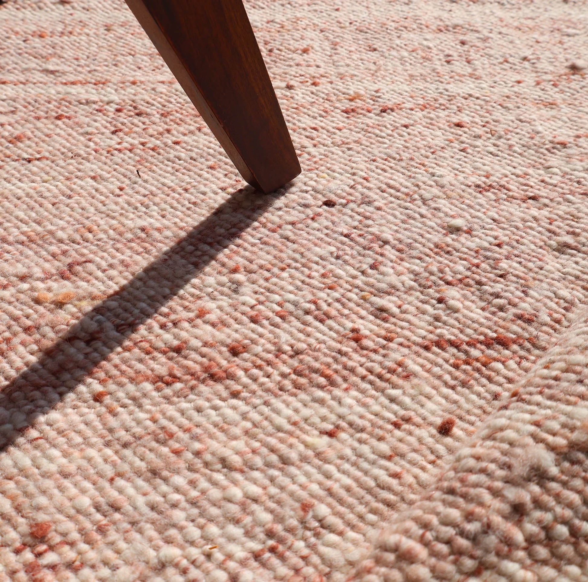 Sunlight highlights the woven texture and gentle ridges of the RAMSHA HOME Dusty Rose Artisan Flatweave Wool Rug (SRN-042), as a wooden furniture leg casts a shadow across its neutral, pinkish-beige fibers with subtle reddish speckles.
