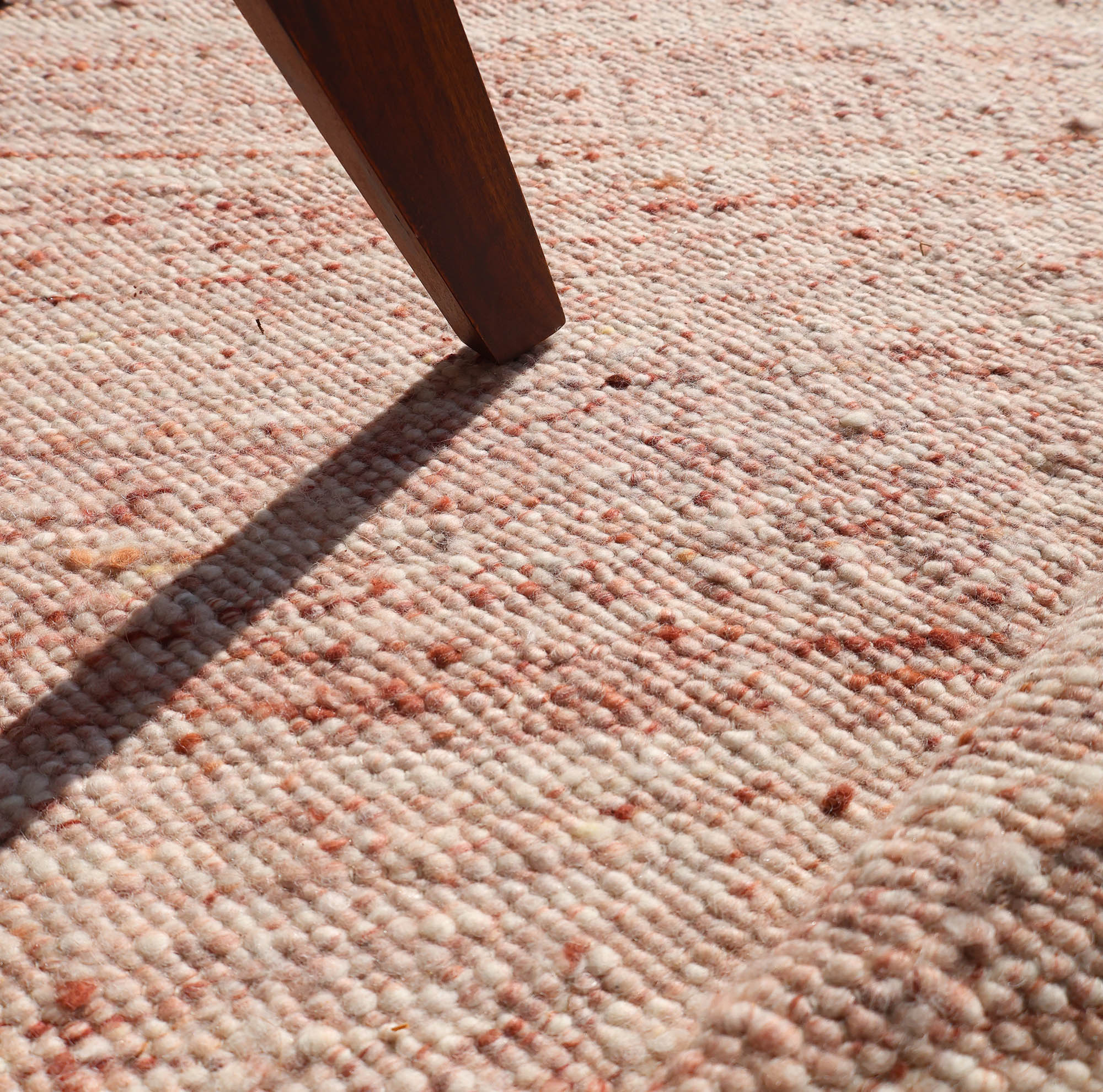 Sunlight highlights the woven texture and gentle ridges of the RAMSHA HOME Dusty Rose Artisan Flatweave Wool Rug (SRN-042), as a wooden furniture leg casts a shadow across its neutral, pinkish-beige fibers with subtle reddish speckles.