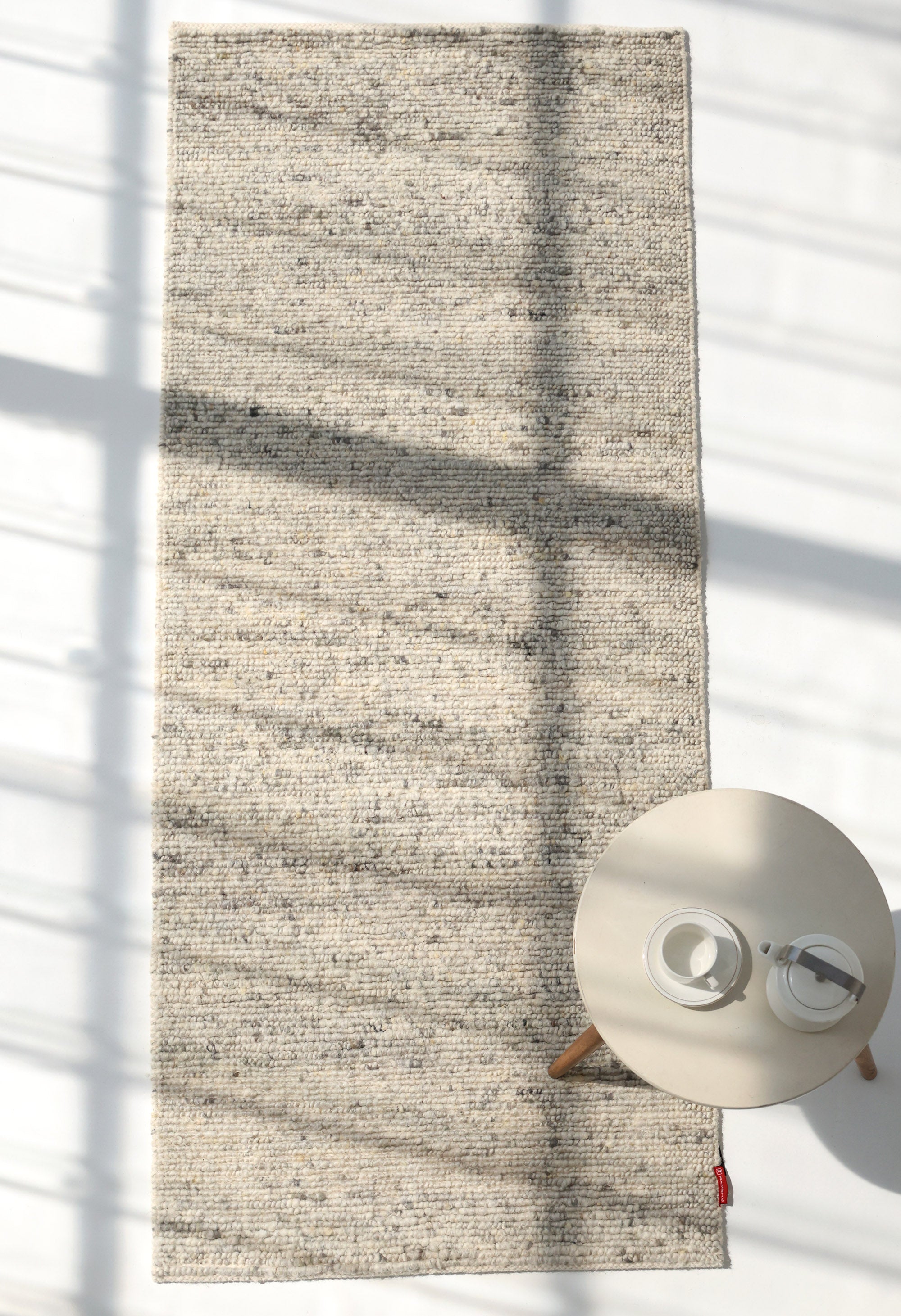 The Soft Terra Flatweave Wool Rug- SRN-043 by RAMSHA HOME, in light beige, lies on a white floor with windowpane shadows. A small round table with two white cups sits at the bottom right of this neutral-toned handmade rug.