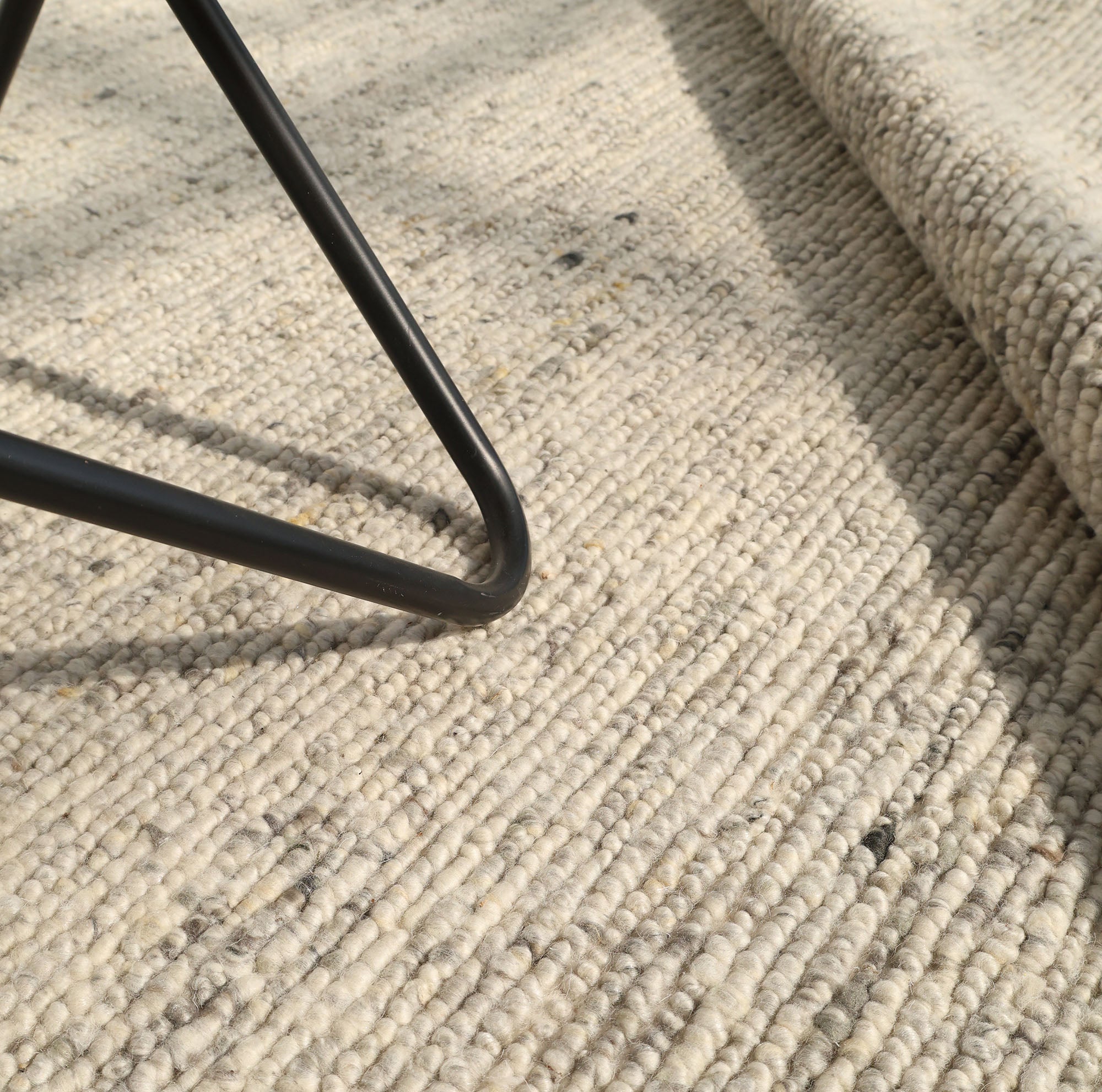 Close-up of the RAMSHA HOME Soft Terra Flatweave Wool Rug (SRN-043) in beige, featuring its textured handmade design under a metal furniture leg with part rolled up. Sunlight casts soft shadows, emphasizing traditional craftsmanship.