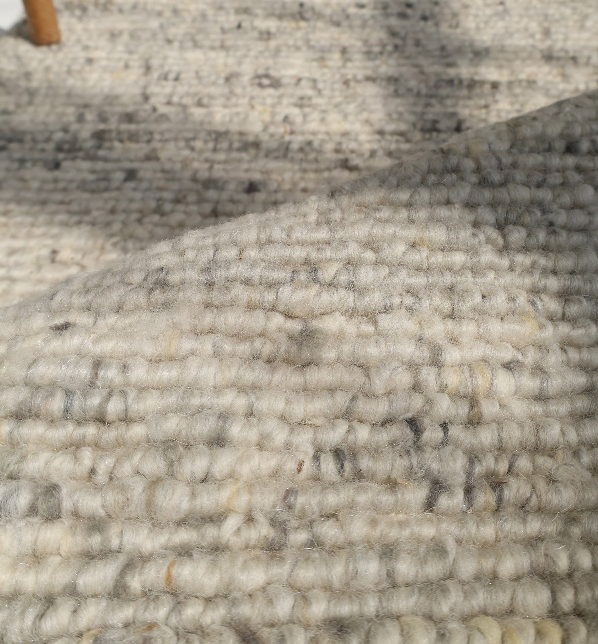 Close-up of the Soft Terra Flatweave Wool Rug- SRN-043 by RAMSHA HOME, showcasing thick, looped beige and gray fibers for an organic texture. Natural light highlights a shadow and part of a wooden furniture leg in the background.