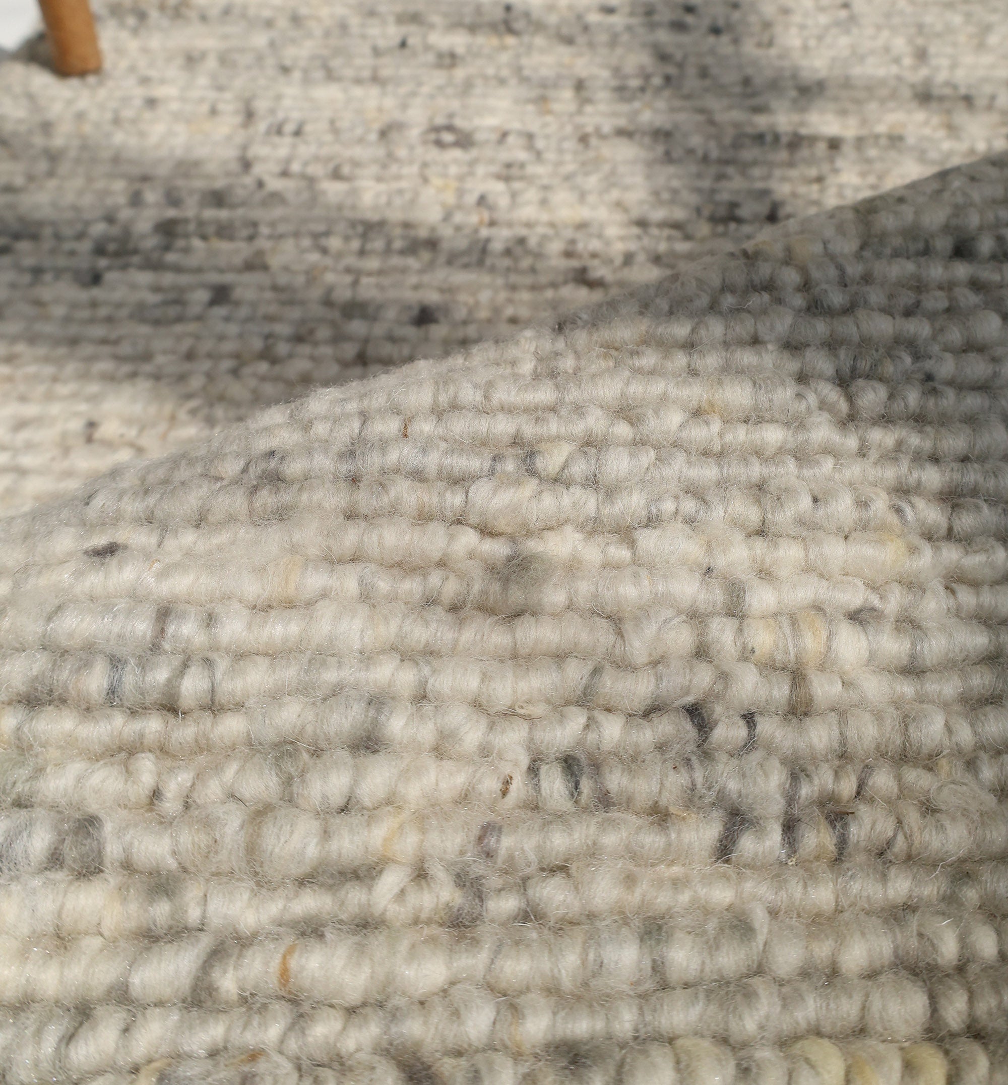Close-up of the Soft Terra Flatweave Wool Rug- SRN-043 by RAMSHA HOME, showcasing thick, looped beige and gray fibers for an organic texture. Natural light highlights a shadow and part of a wooden furniture leg in the background.