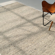 The Soft Terra Flatweave Wool Rug (SRN-043) by RAMSHA HOME adds textured charm to a sunlit room, its neutral tones accented by dancing window shadows, a brown leather butterfly chair, and a round white side table in the upper right corner.