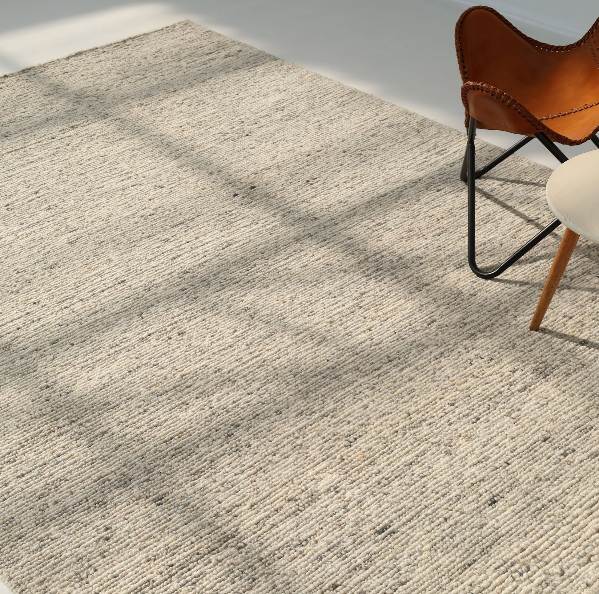 The Soft Terra Flatweave Wool Rug (SRN-043) by RAMSHA HOME adds textured charm to a sunlit room, its neutral tones accented by dancing window shadows, a brown leather butterfly chair, and a round white side table in the upper right corner.