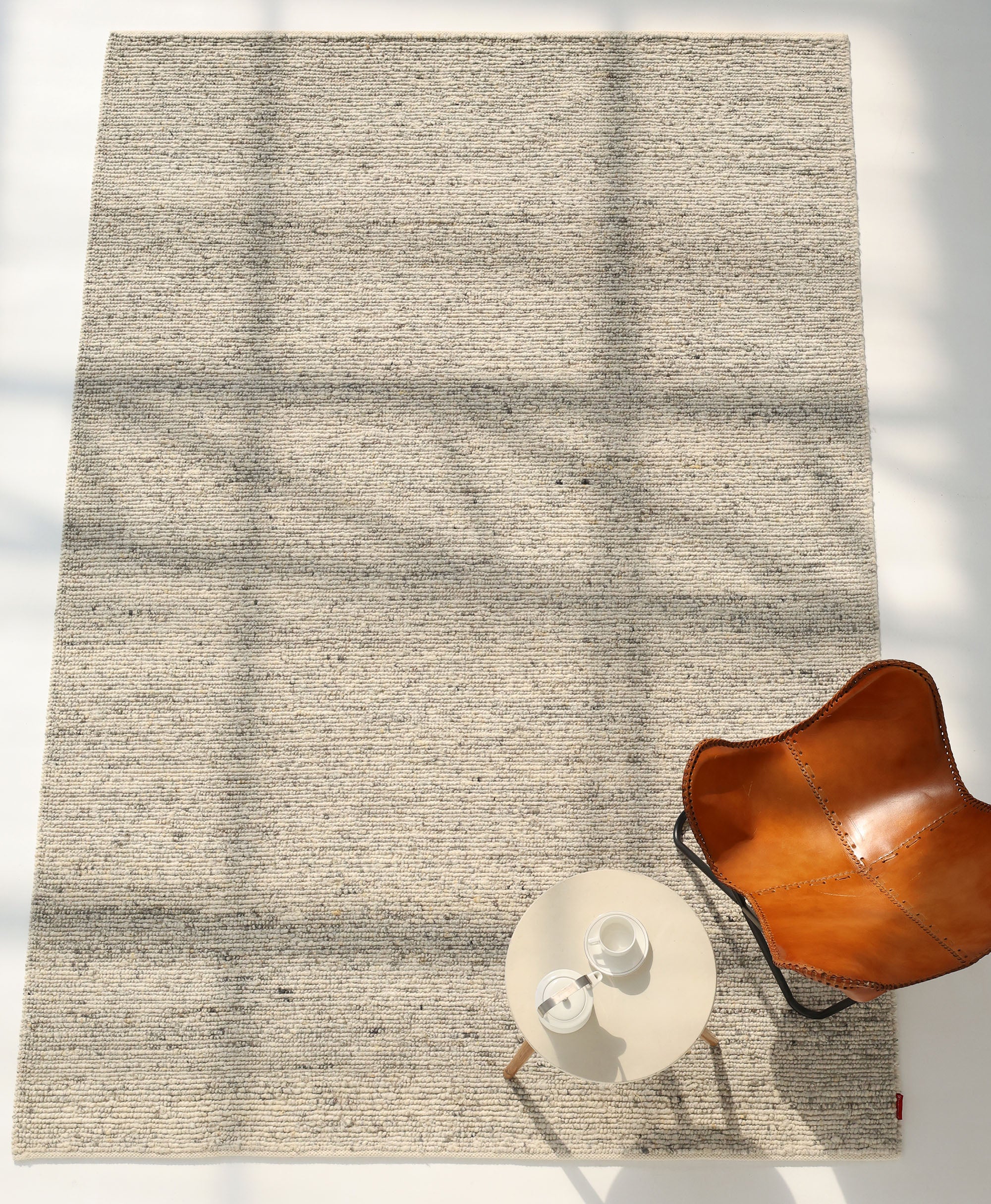 The Soft Terra Flatweave Wool Rug- SRN-043 by RAMSHA HOME features neutral tones and horizontal lines, paired with a tan leather chair and a round side table with tea ware. Soft shadows highlight its traditional craftsmanship in this minimalist space.