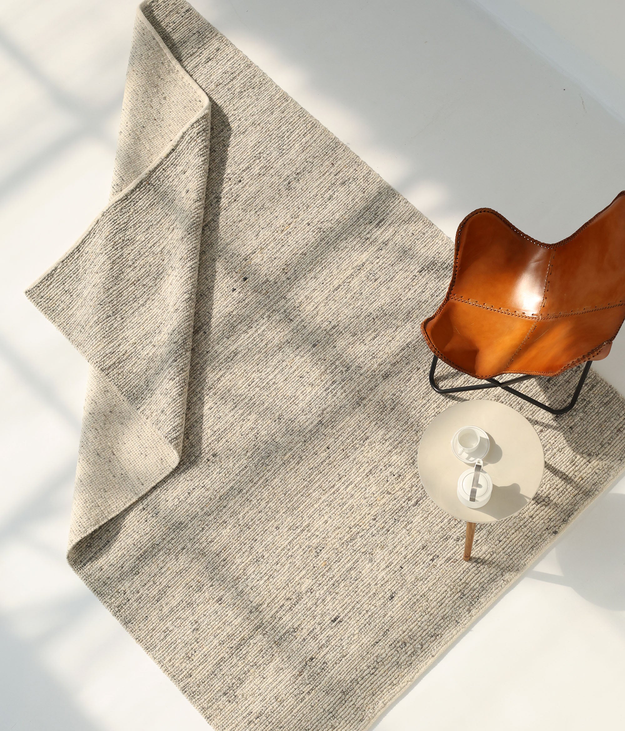 The Soft Terra Flatweave Wool Rug (SRN-043) by RAMSHA HOME lies partially folded on a white floor beside a round side table with a cup and saucer and a brown leather chair, as sunlight casts soft shadows across the scene.