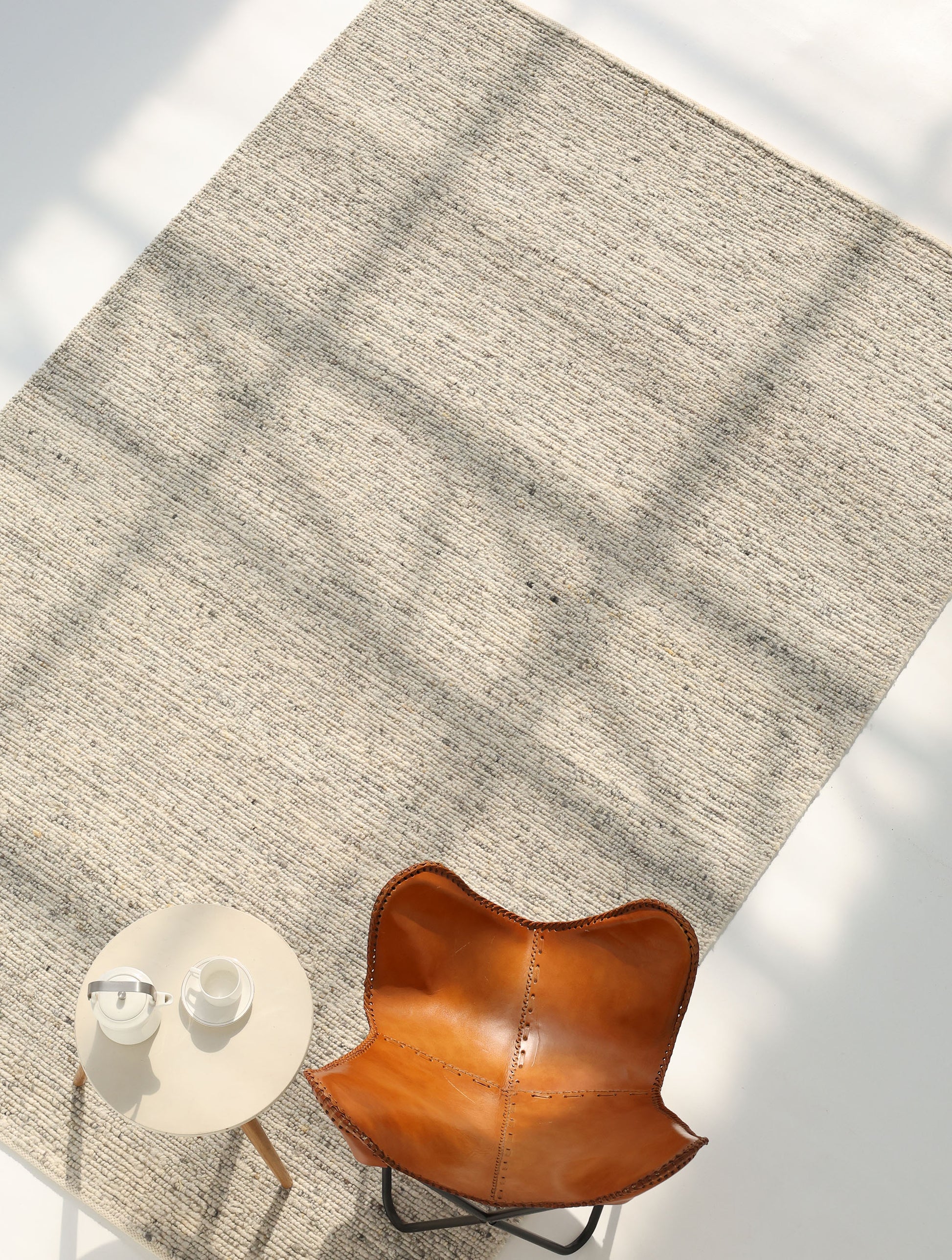 A tan leather chair sits beside a small round table with water and a carafe, atop the RAMSHA HOME Soft Terra Flatweave Wool Rug (SRN-043) in a sunlit, minimal space that highlights traditional craftsmanship.