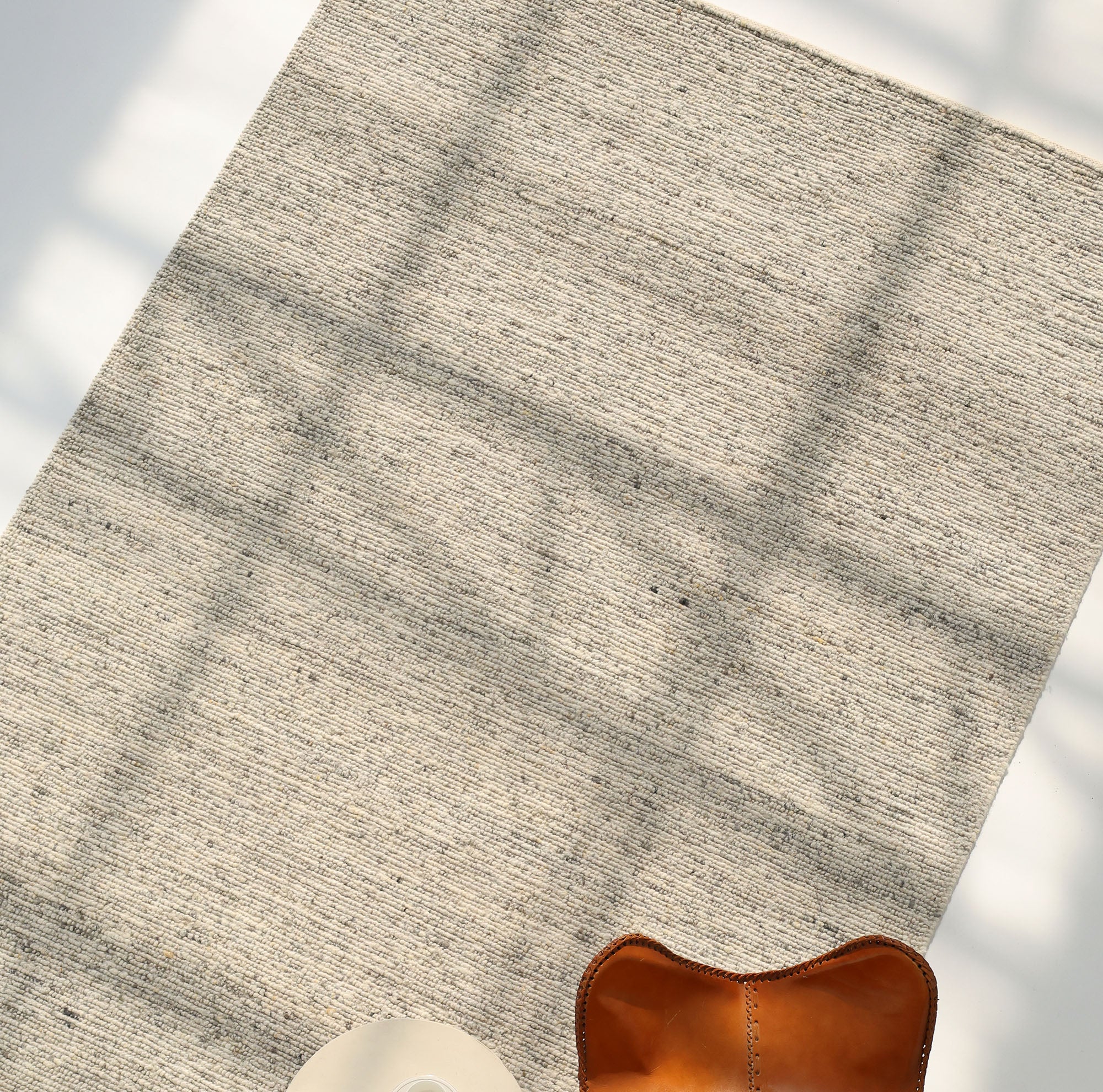 The Soft Terra Flatweave Wool Rug (SRN-043) by RAMSHA HOME, crafted with traditional techniques in a neutral hue, lies on the floor amid window-cast shadows, partially revealing a brown leather chair and the edge of a round white table.
