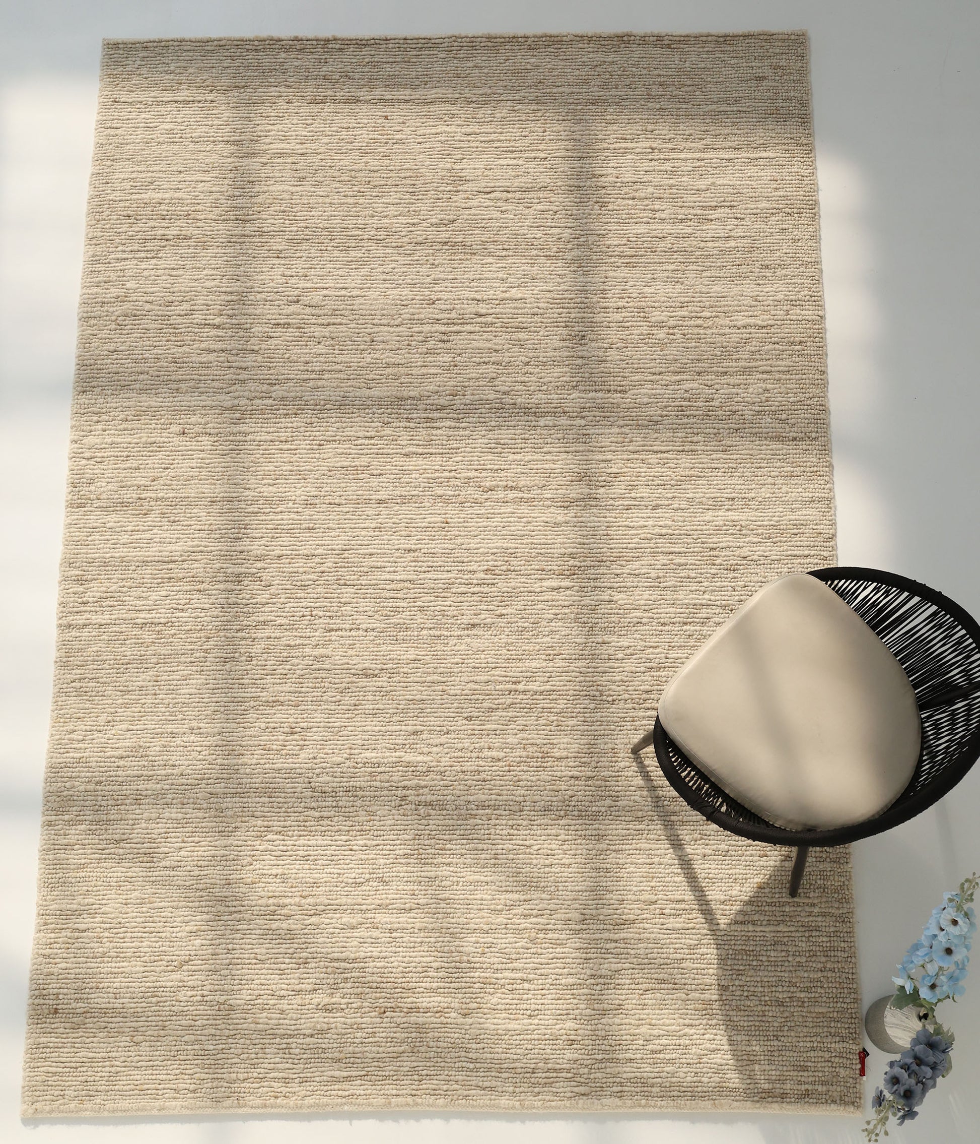 The RAMSHA HOME Earthy Blush Contemporary Flatweave Rug (SRN-044) is spread on the floor with patterns of light and shadow. A black chair with a light cushion sits nearby, next to a small bouquet of blue flowers.