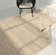 The RAMSHA HOME Earthy Blush Contemporary Flatweave Rug (SRN-044) in light beige is partially folded on a white floor, with a black woven chair and beige cushion nearby in a bright room.