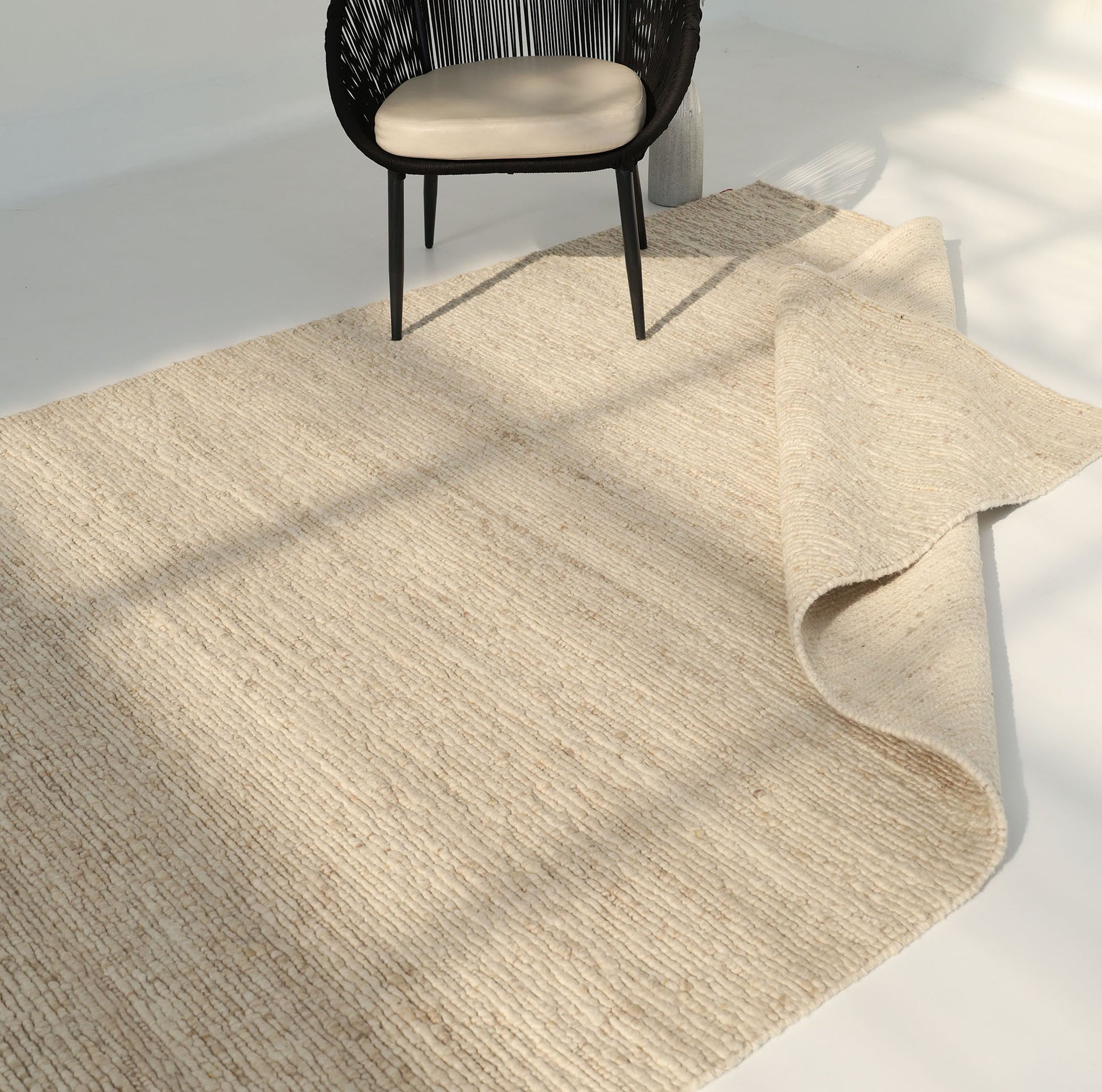 The RAMSHA HOME Earthy Blush Contemporary Flatweave Rug (SRN-044) in light beige is partially folded on a white floor, with a black woven chair and beige cushion nearby in a bright room.