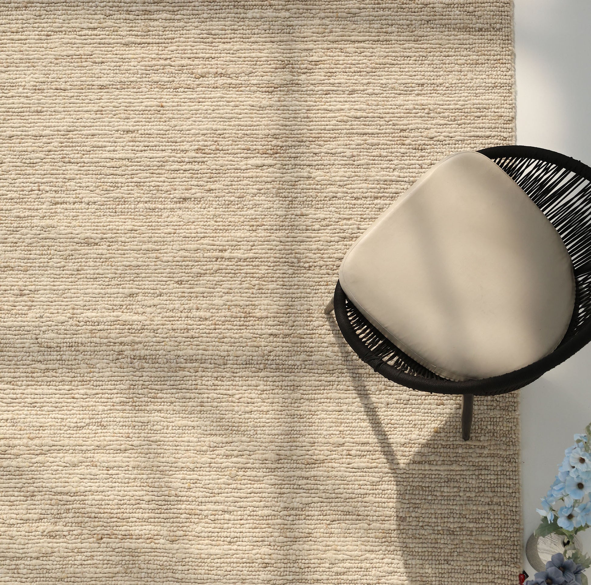 The Earthy Blush Contemporary Flatweave Rug by RAMSHA HOME covers the floor. A black wicker chair with a light beige cushion sits in the top right, while light blue flowers are at the bottom right. Sunlight casts soft shadows across the room.