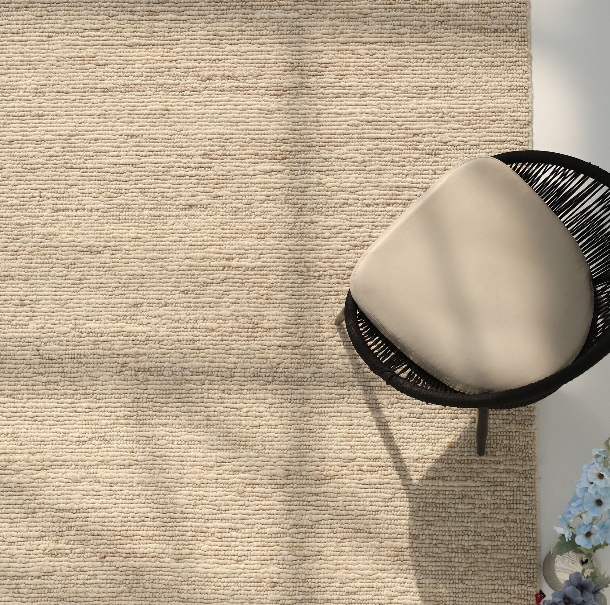 The Earthy Blush Contemporary Flatweave Rug by RAMSHA HOME covers the floor. A black wicker chair with a light beige cushion sits in the top right, while light blue flowers are at the bottom right. Sunlight casts soft shadows across the room.