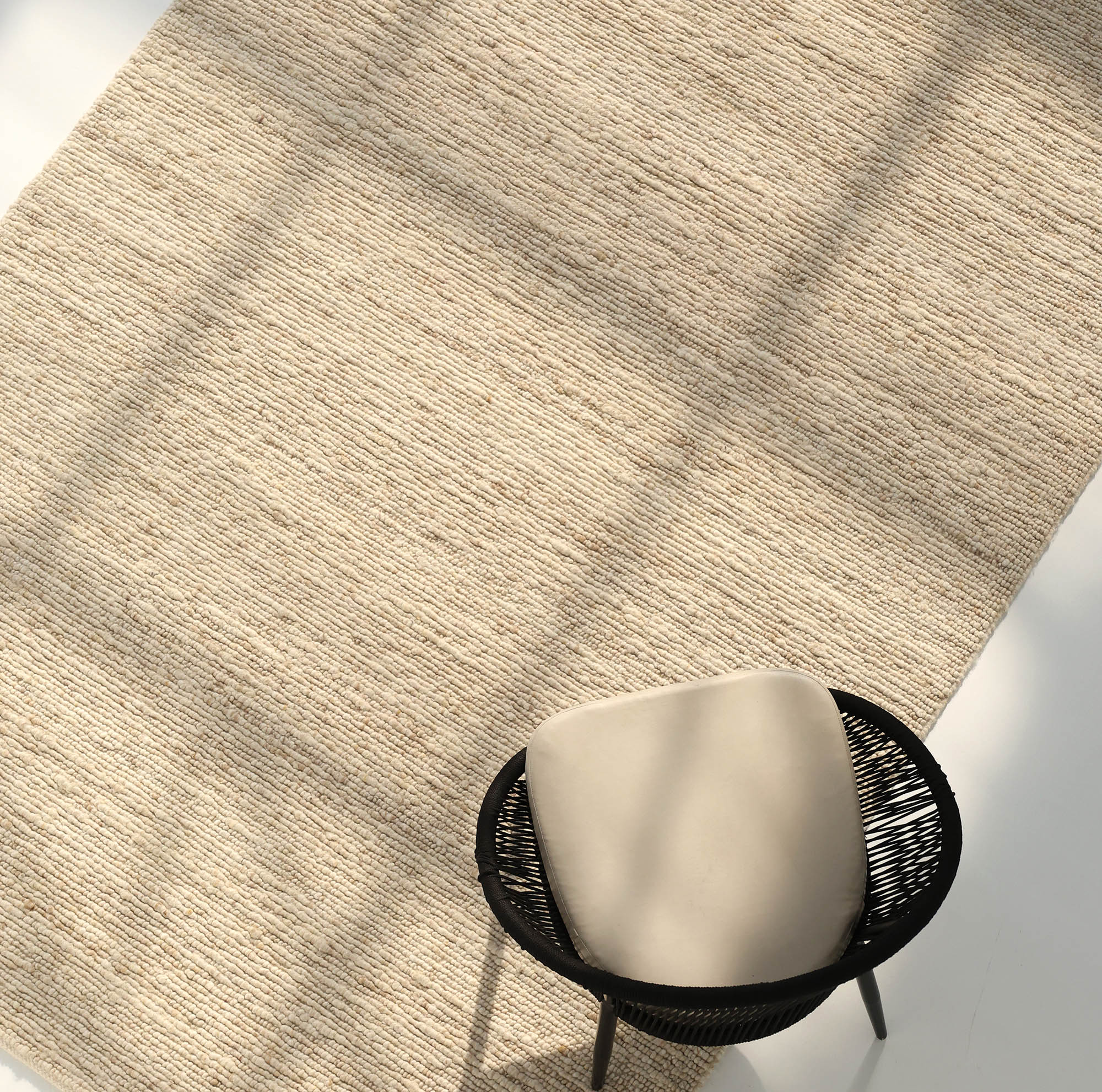Sunlight streams across the RAMSHA HOME Earthy Blush Contemporary Flatweave Rug (SRN-044), casting soft shadows. A black wicker chair with a light cushion sits partially on the rug, accentuating its traditional craftsmanship.