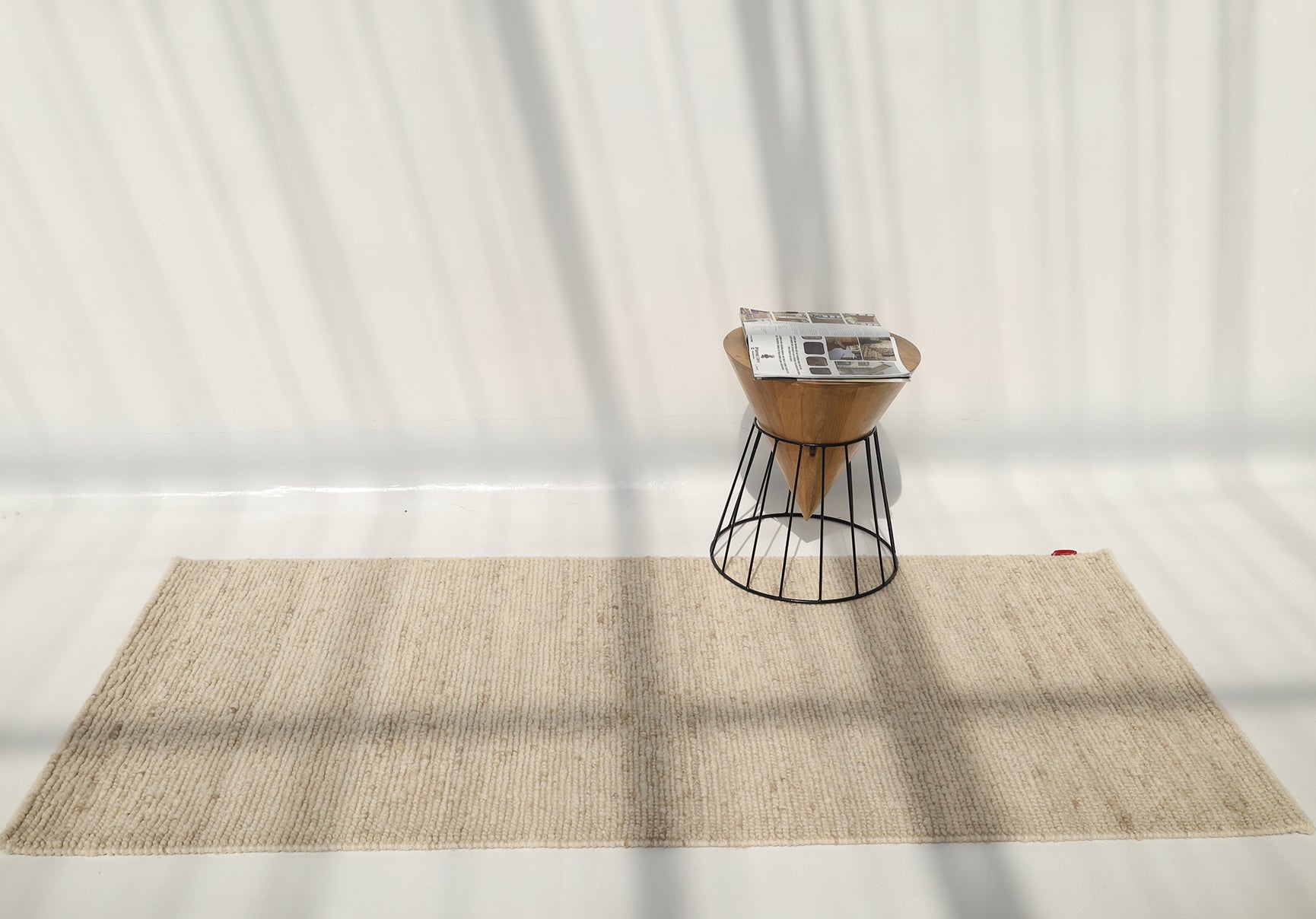 A small wooden stool with a metal base holds newspapers and sits on the RAMSHA HOME Earthy Blush Contemporary Flatweave Rug (SRN-044) in a sunny room, sunlight casting shadows across the white wall and floor.