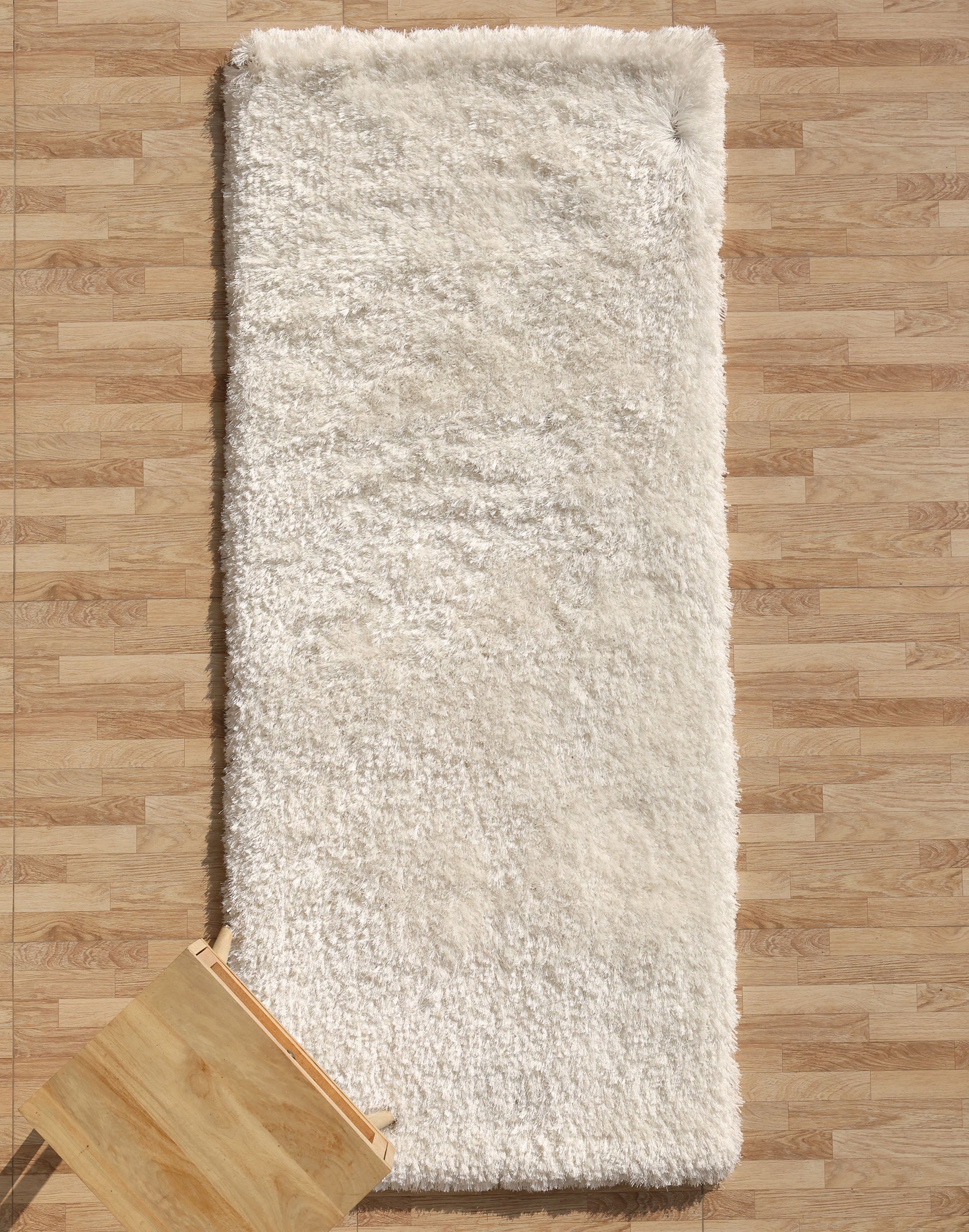 The RAMSHA HOME White Color Plush Shaggy Area Rug - SG 040 rests on a light wood floor, its plush fibers creating a cozy vibe. A small wooden stool is partially visible in the lower left corner.